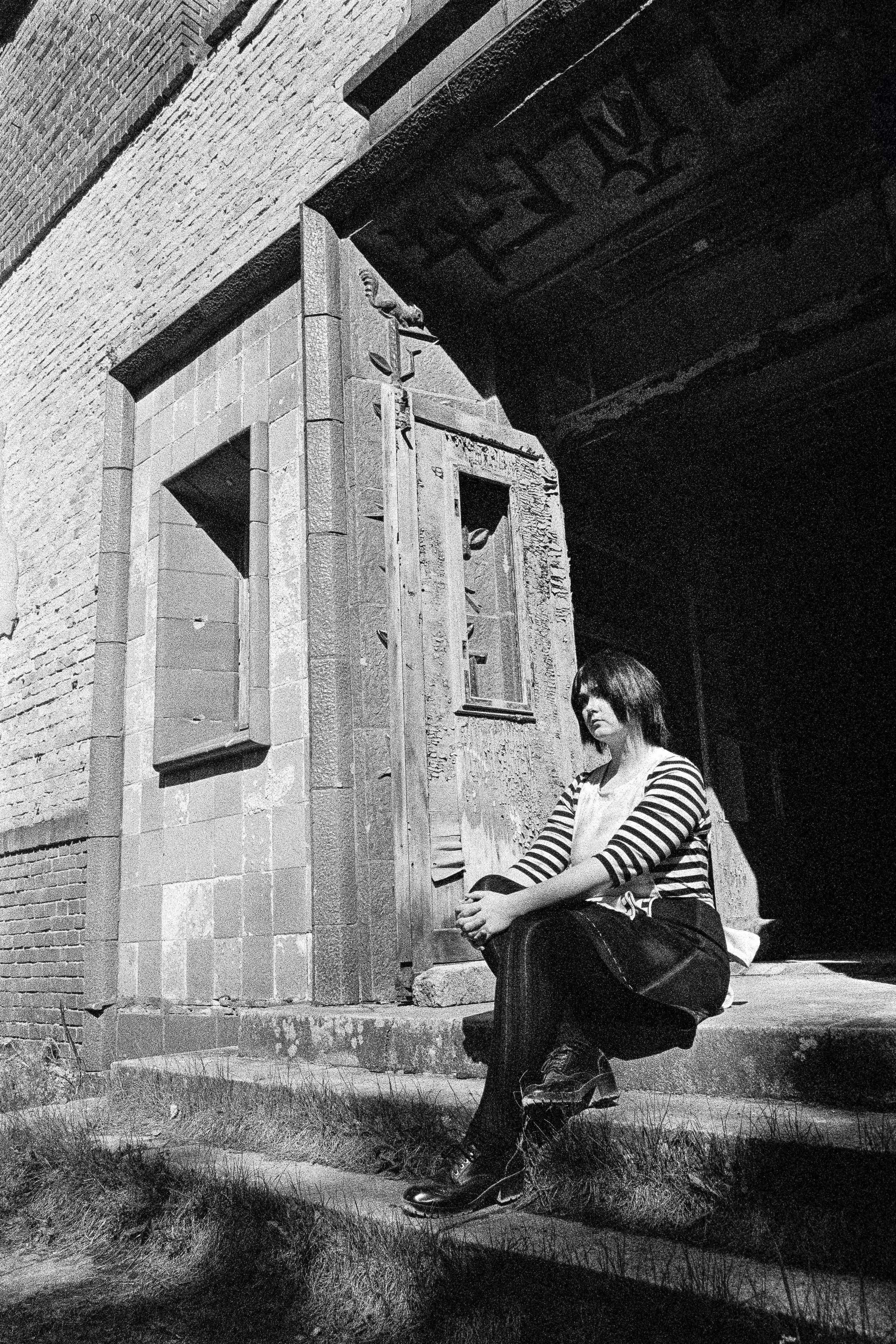 Schwarz-Weiß-Fotografie einer jungen Frau mit schwarzen Haaren, die auf den Stufen vor einer alten Steinfassade sitzt, mit einem Gebäude im Hintergrund.