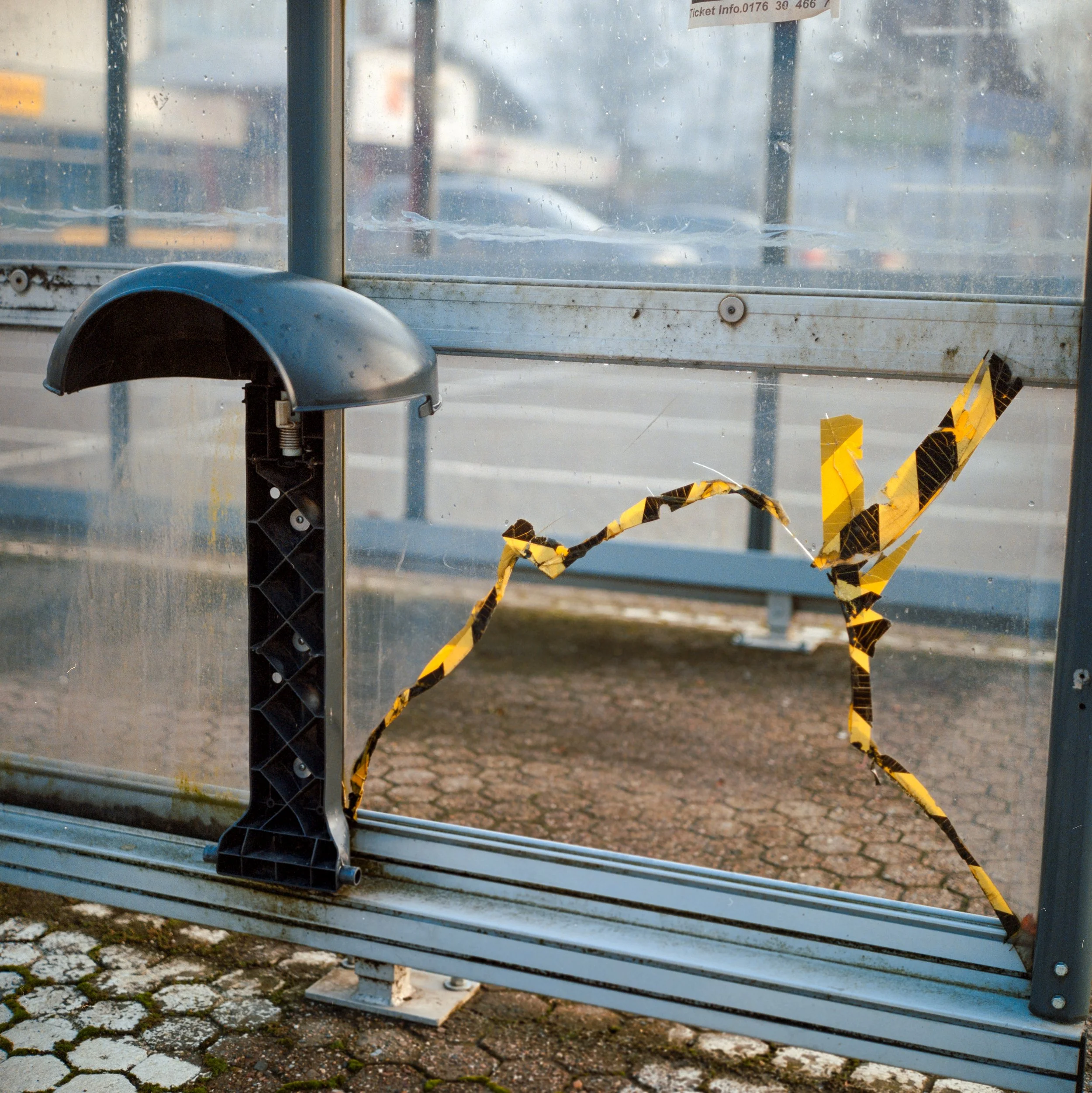 Eine kaputte Bushaltestelle mit zerstörtem Glas und abgesplittertem schwarzem Plastik. Das Gelb-Schwarz-Klebeband klebt markiert die Risse im Glas.