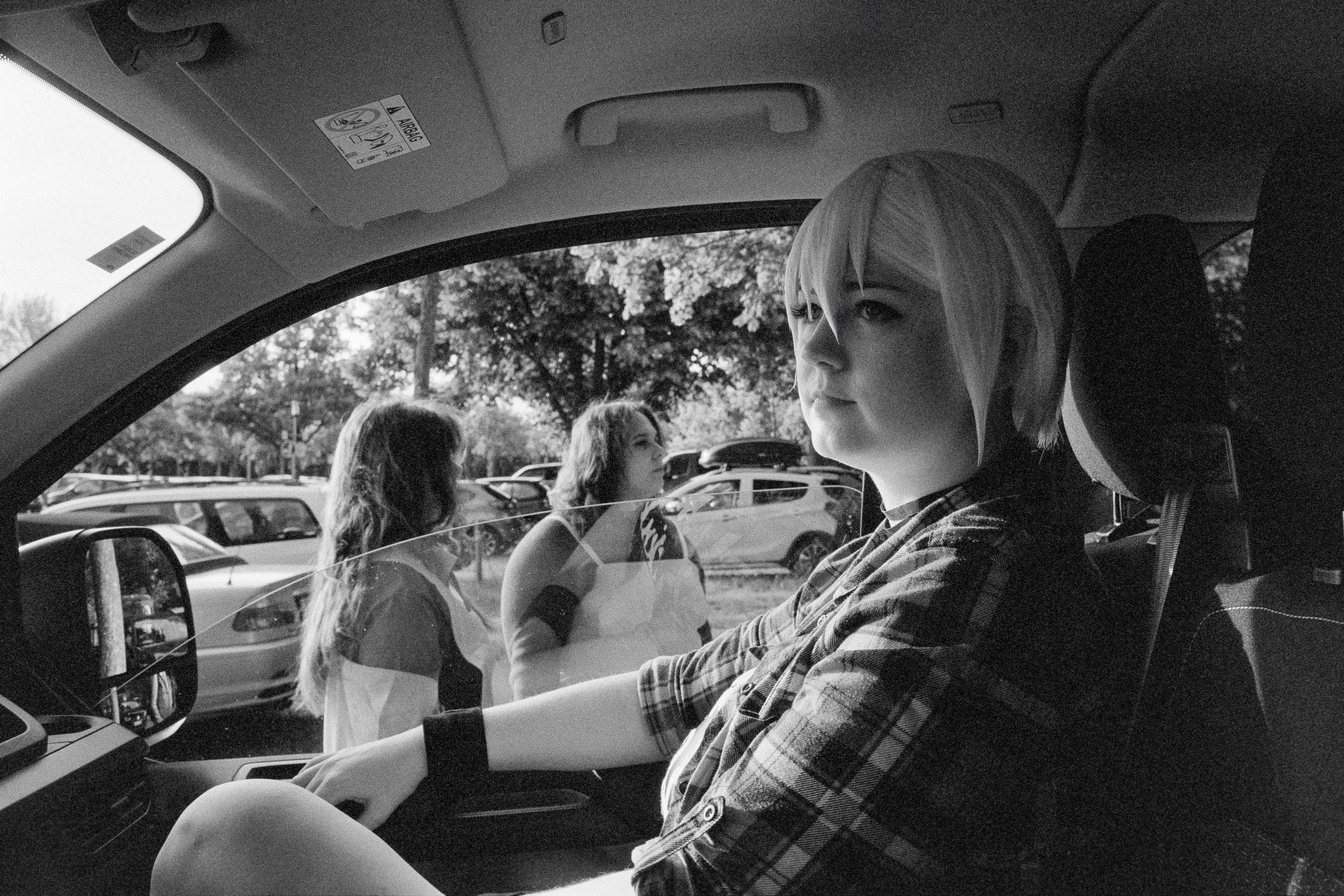 Schwarz-Weiß-Fotografie eines Mädchens mit blondem Haar, das im Auto sitzt, während draußen zwei andere Mädchen an einer Parkplatzzone vorbeigehen.
