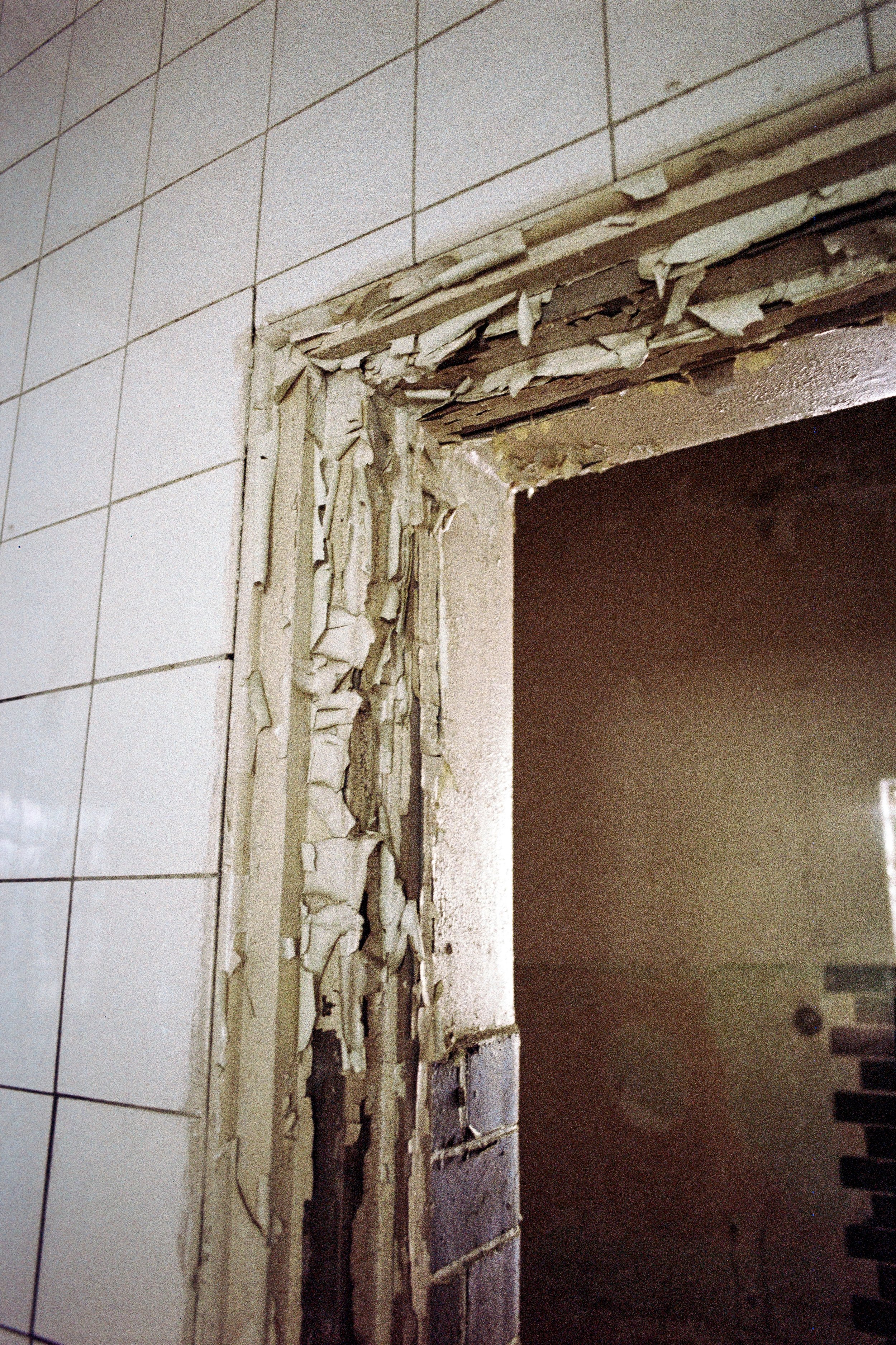 Abgeblätterte Farbe und beschädigter Türrahmen in einem Raum mit weißen Wandfliesen.