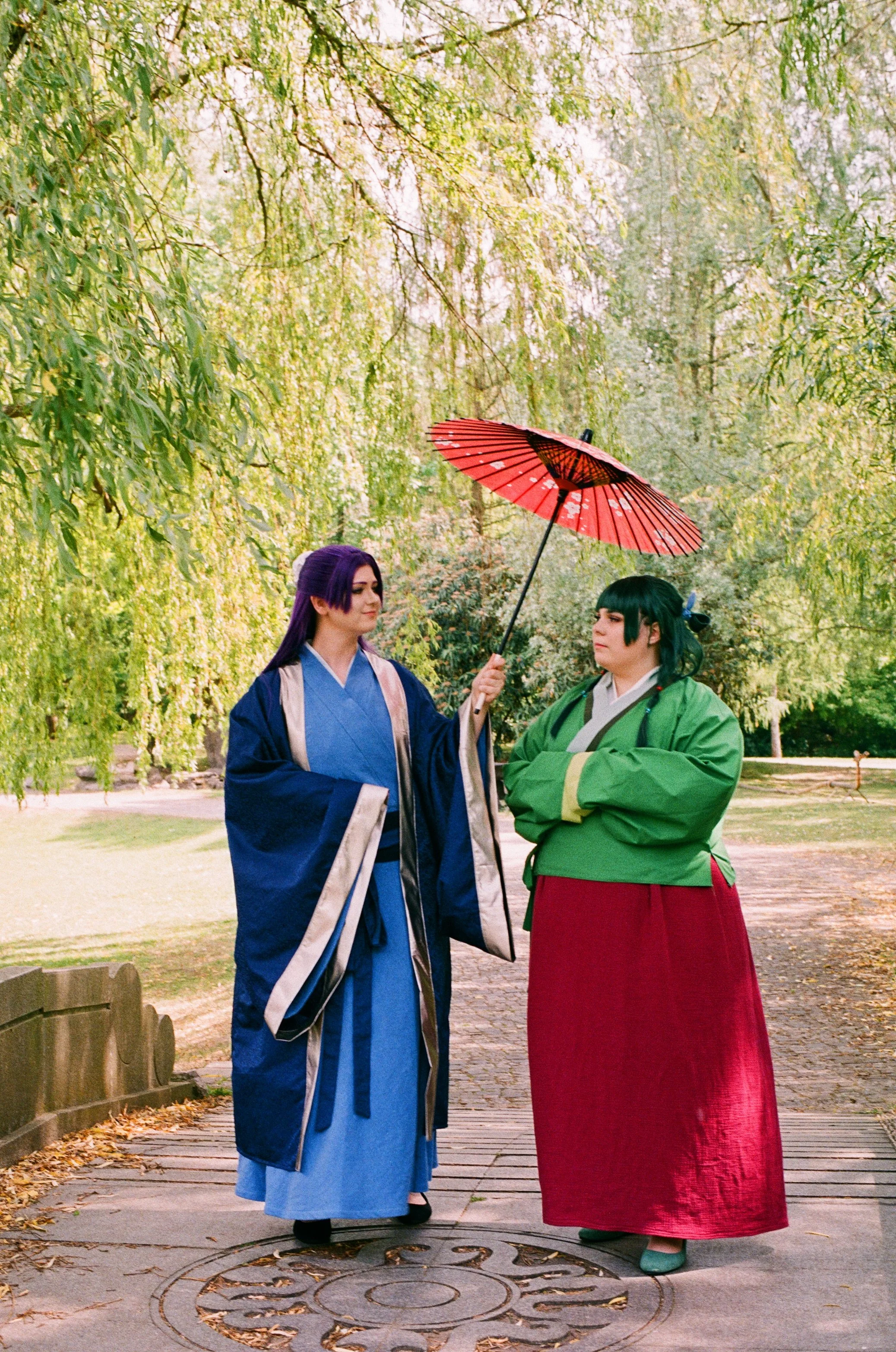 Zwei Personen in traditioneller japanischer Kleidung stehen im Park, eine hält einen roten Regenschirm über die andere.