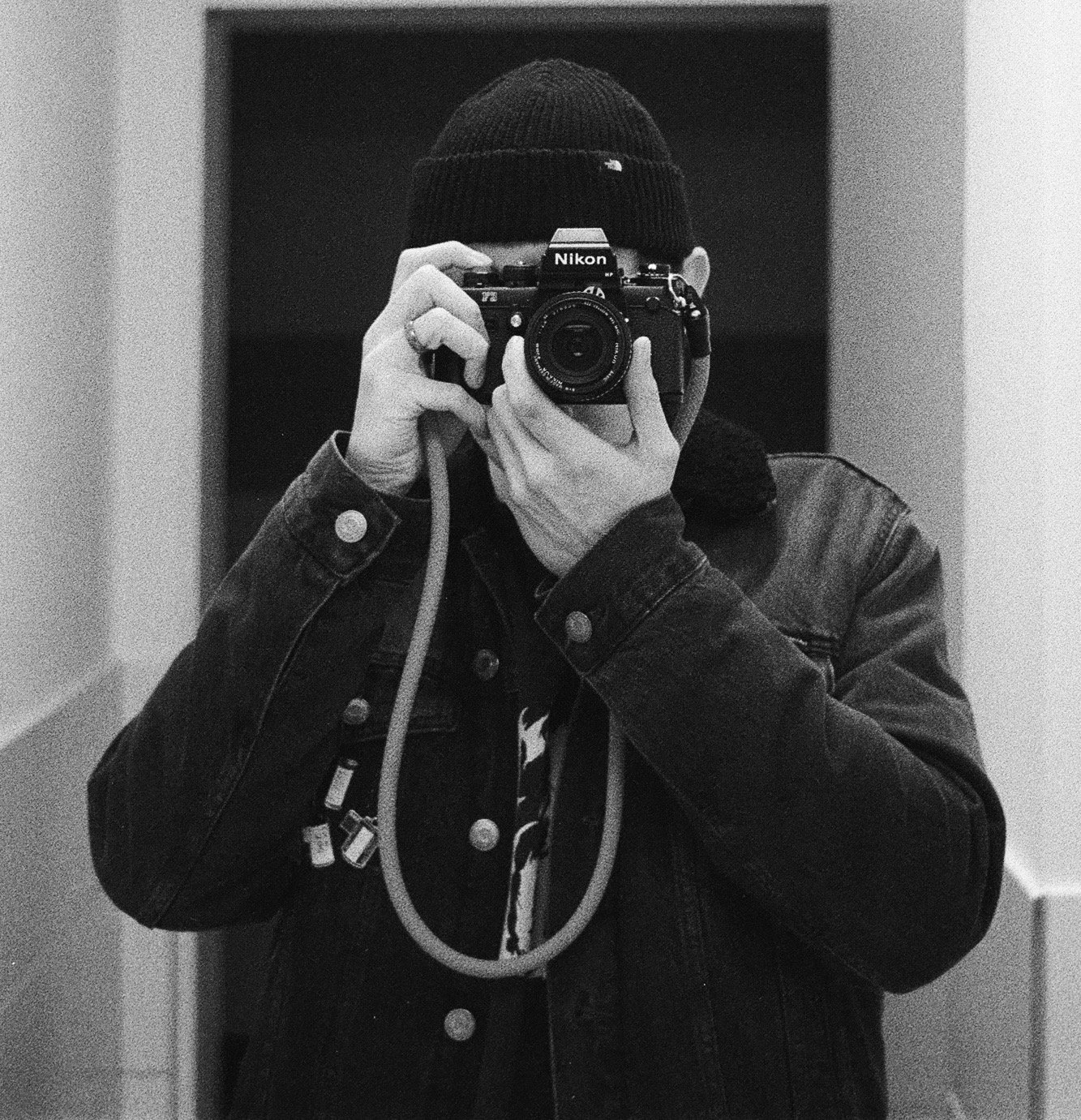 Schwarz-Weiß-Foto einer Person in dunkler Jacke und Mütze, die eine Kamera aufnimmt, im Spiegel sichtbar.