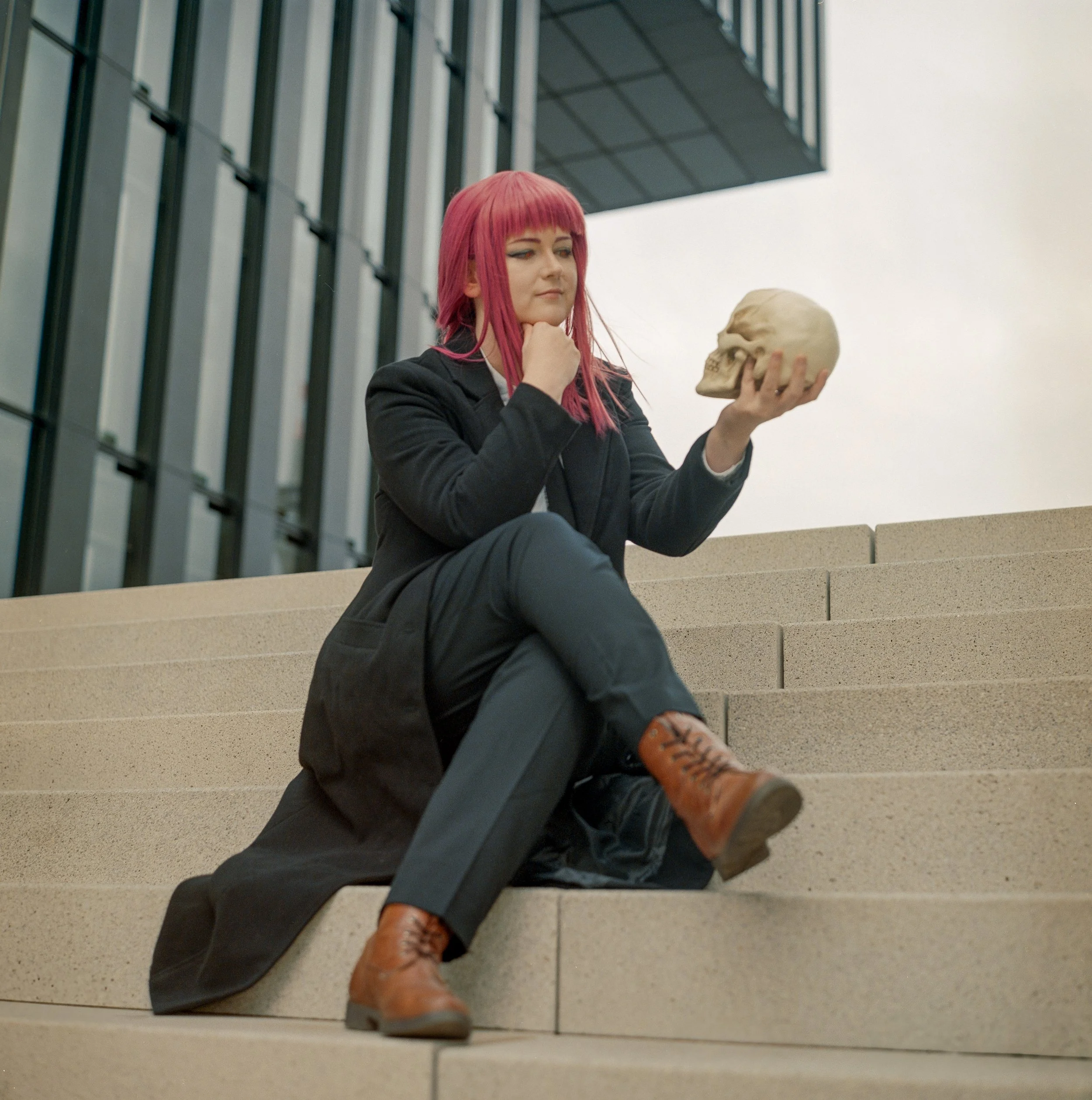 Junge Frau mit pinkem Haar sitzt auf Treppe, hält einen menschlichen Schädel in der Hand und sieht nachdenklich aus, vor einem modernen Gebäude.