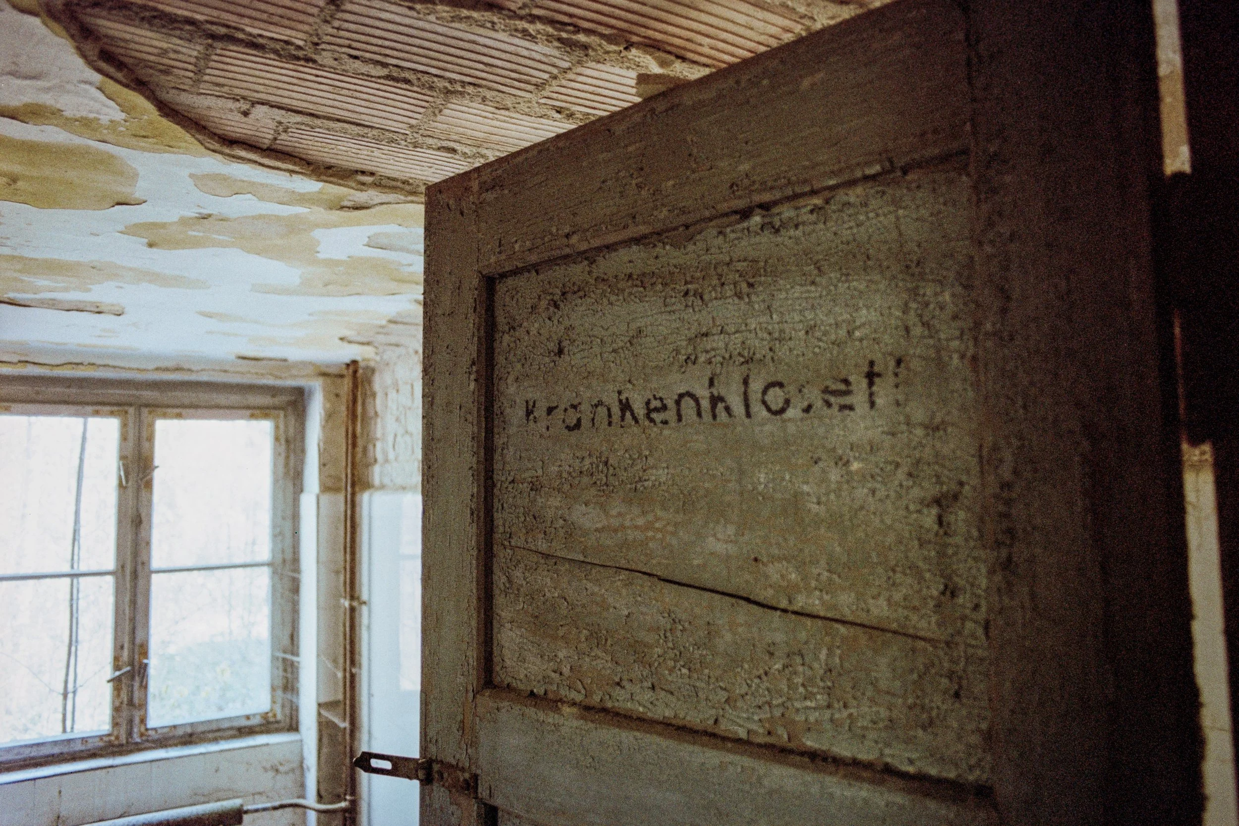 Alte, verbeulte Tür in einem Raum mit abblätternder Deckenfarbe und einem schmutzigen Fenster im Hintergrund.