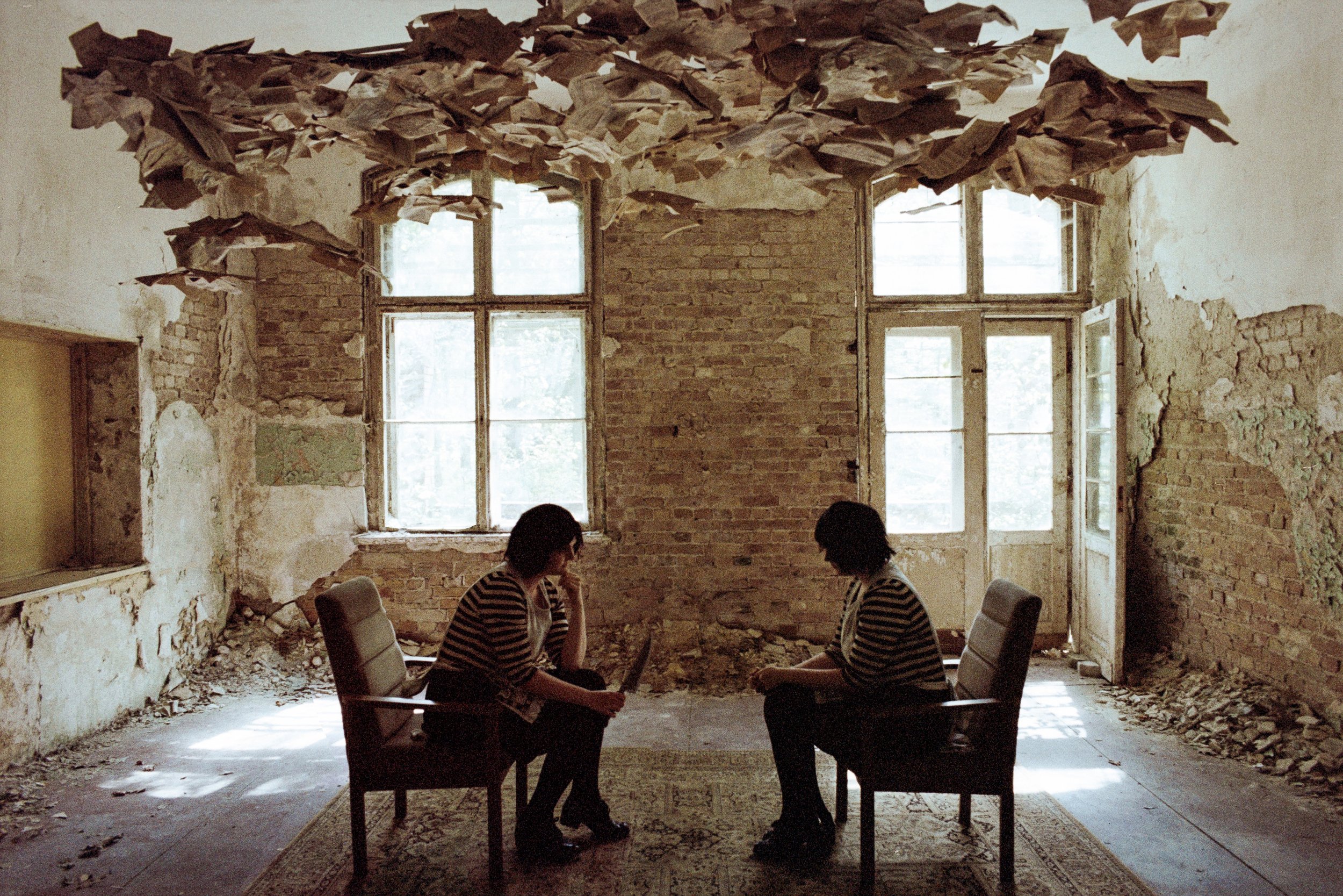 Zwei Frauen sitzen in einem verwohnten, zerfallenden Raum mit großen Fenstern, die wie Spiegel die Natur draußen reflektieren, und Gespräche bei einem Sekt, auf einem alten Teppich sitzend.