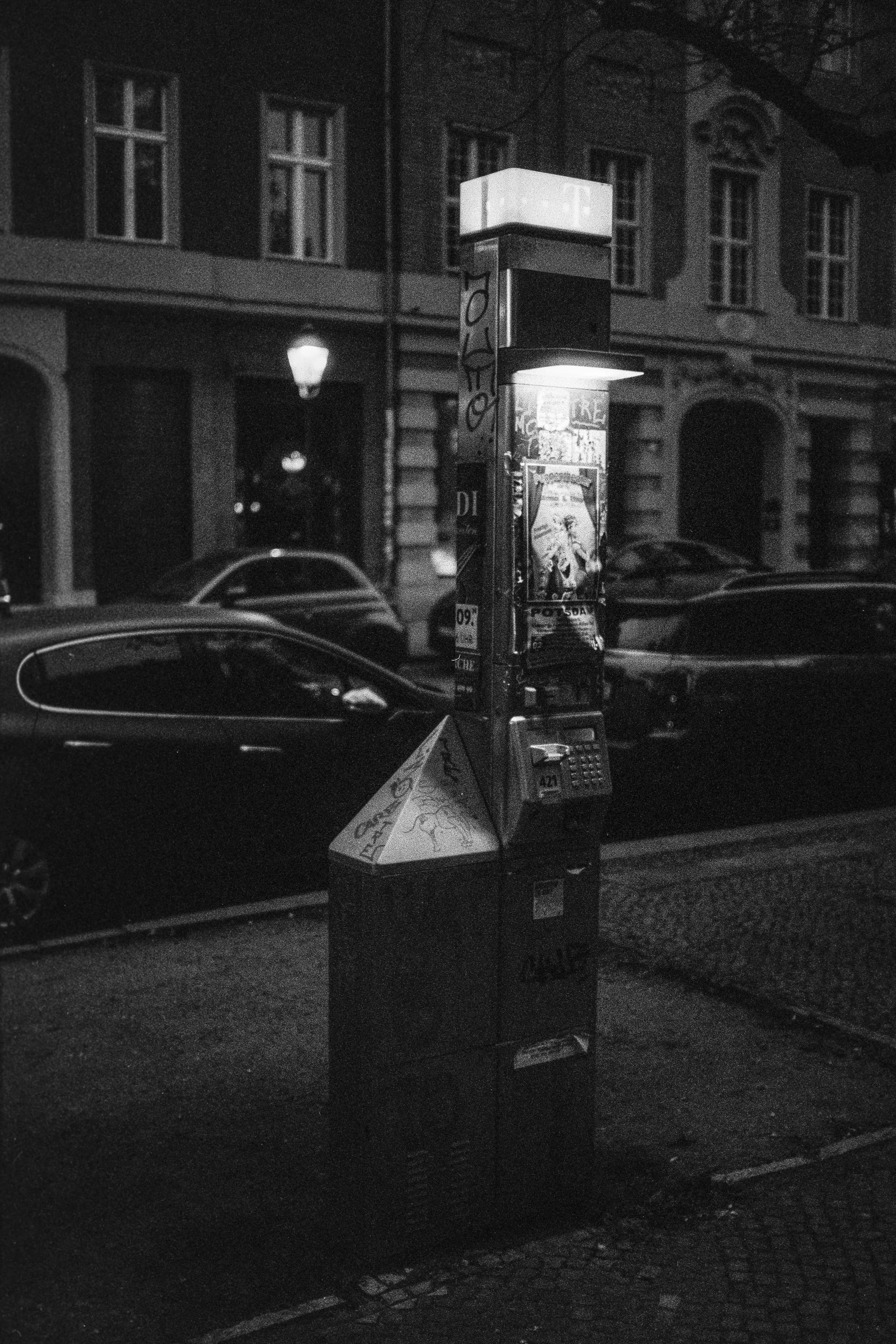 Ein schwarzer, verstaubter Parkscheinautomat auf einer Stadtstraße bei Nacht, umgeben von geparkten Autos und alten Gebäuden.