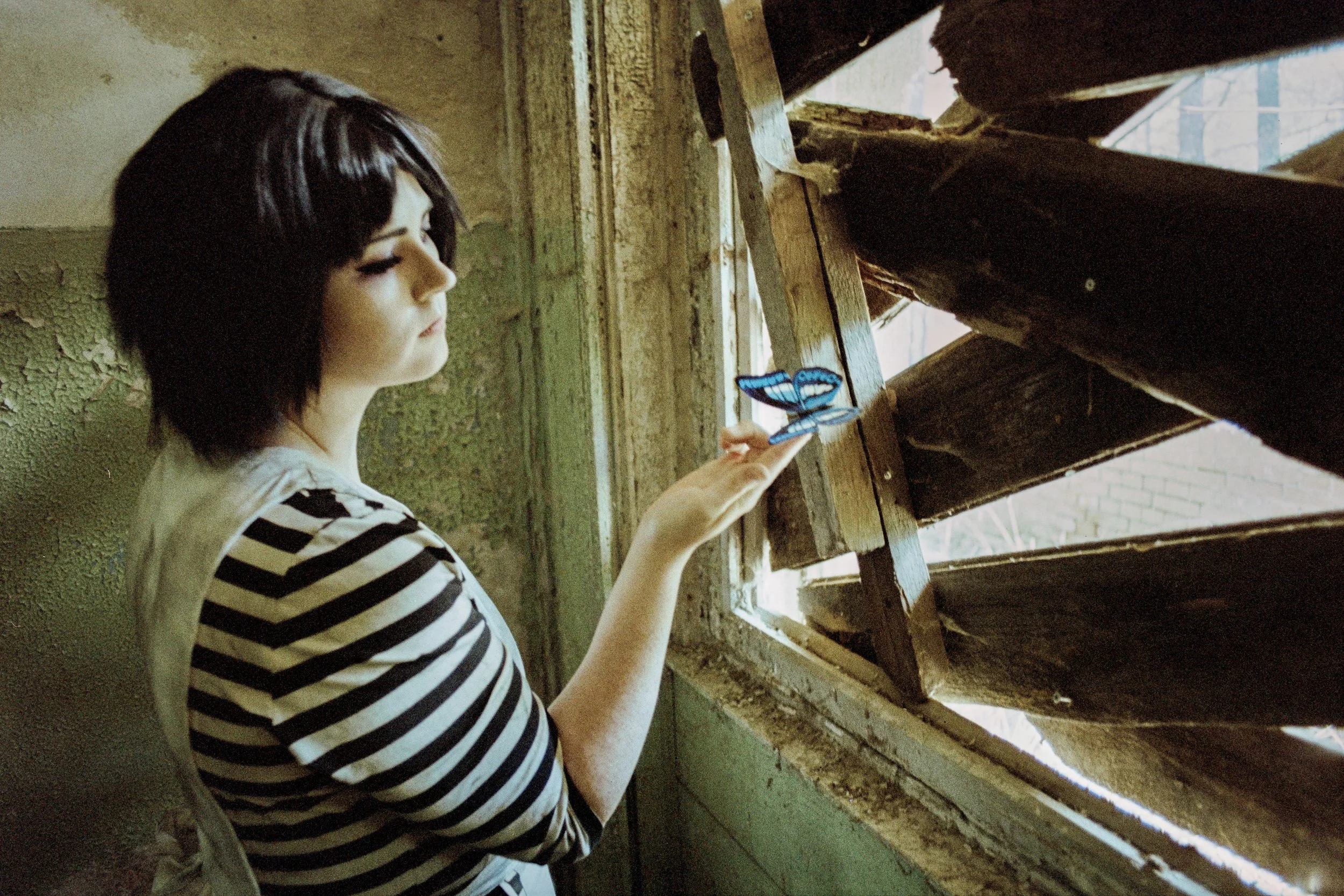 Ein junges Mädchen mit schwarzen Haaren und gestreiftem Pullover hält eine blaue Schmetterlingsdekoration vor einem halb offenen Fenster in einem alten, verfallenen Gebäude.