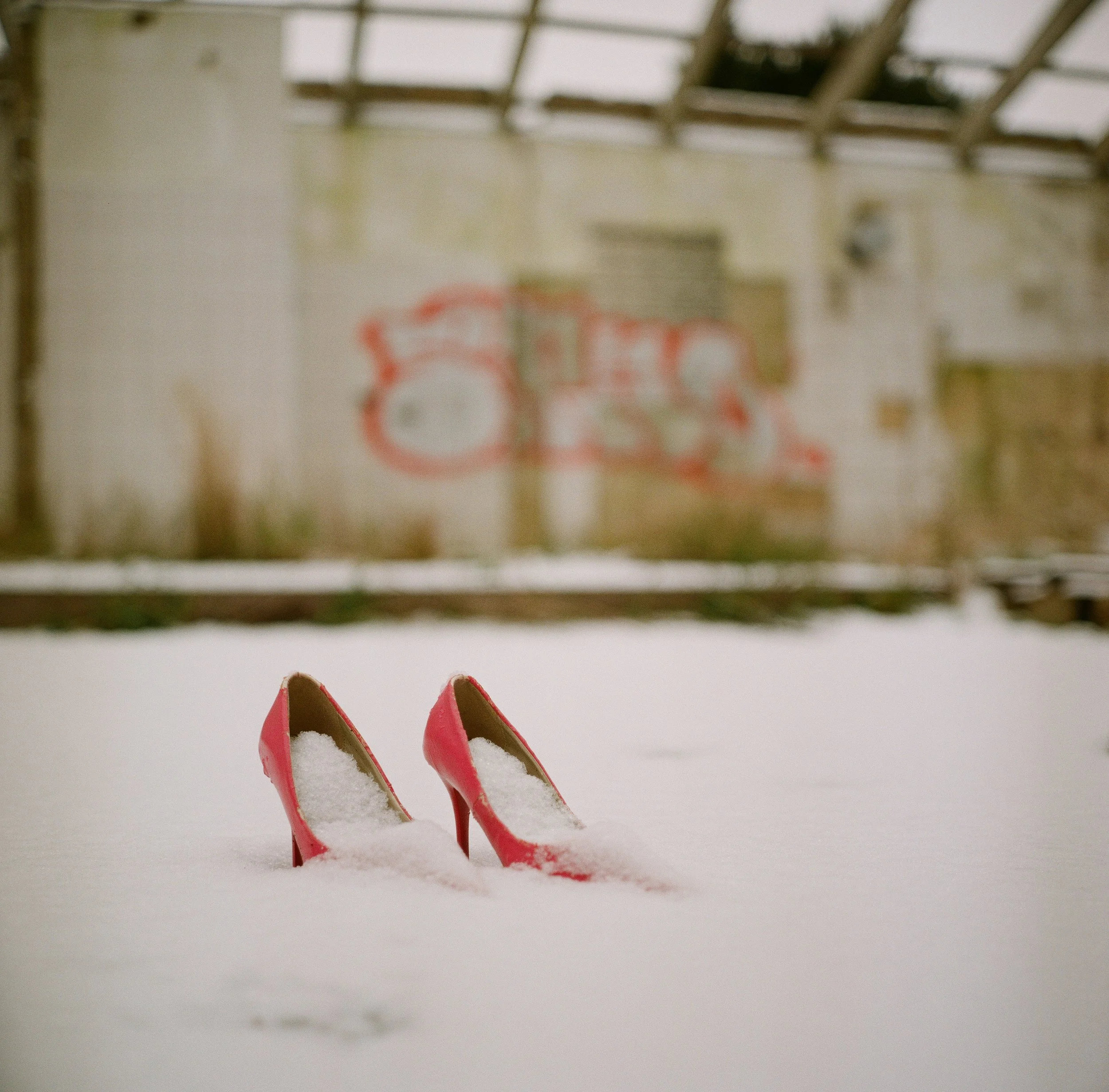 Rote High Heels im Schnee mit einer verfallenen, graffiti-bedeckten Wand im Hintergrund.