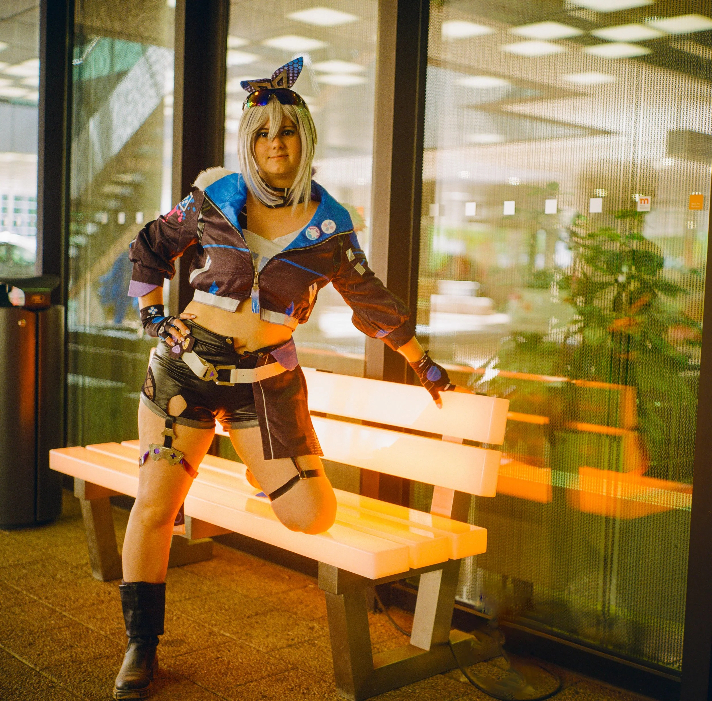 Person in Cosplay-Kostüm steht neben einer Bank in einem Wartebereich, trägt ein buntes, futuristisches Outfit mit Tierohren und Sonnenbrille, im Hintergrund Glasfenster mit Blick nach draußen.