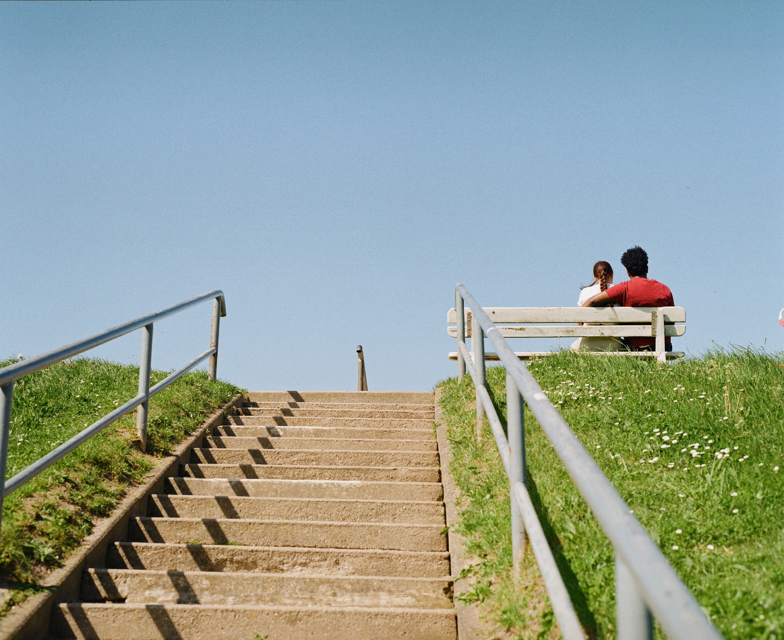 Zwei Menschen, ein Mann und eine Frau, sitzen auf einer Bank am oberen Ende einer Treppe, die nach oben führt, im Freien unter einem blauen Himmel.