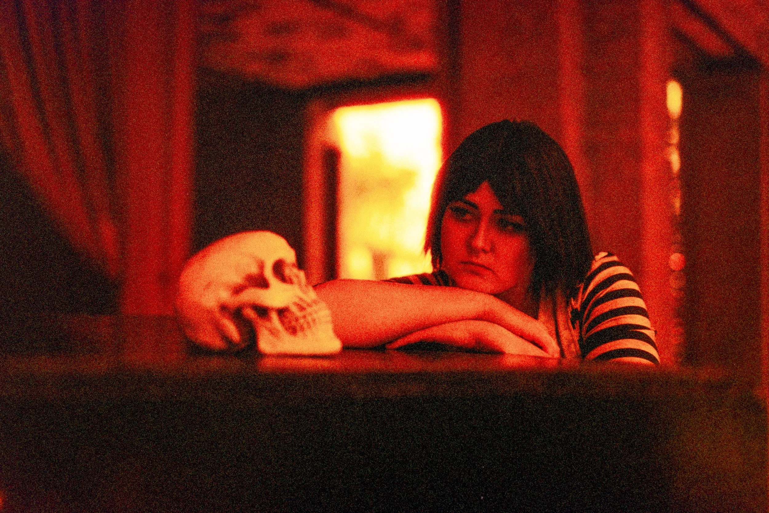 Eine junge Frau mit dunklem, kurzem Haar liegt mit ihren Armen auf einem Tisch und schaut nachdenklich auf einen menschlichen Schädel, der auf dem Tisch liegt, in einem Raum mit rotem Licht.