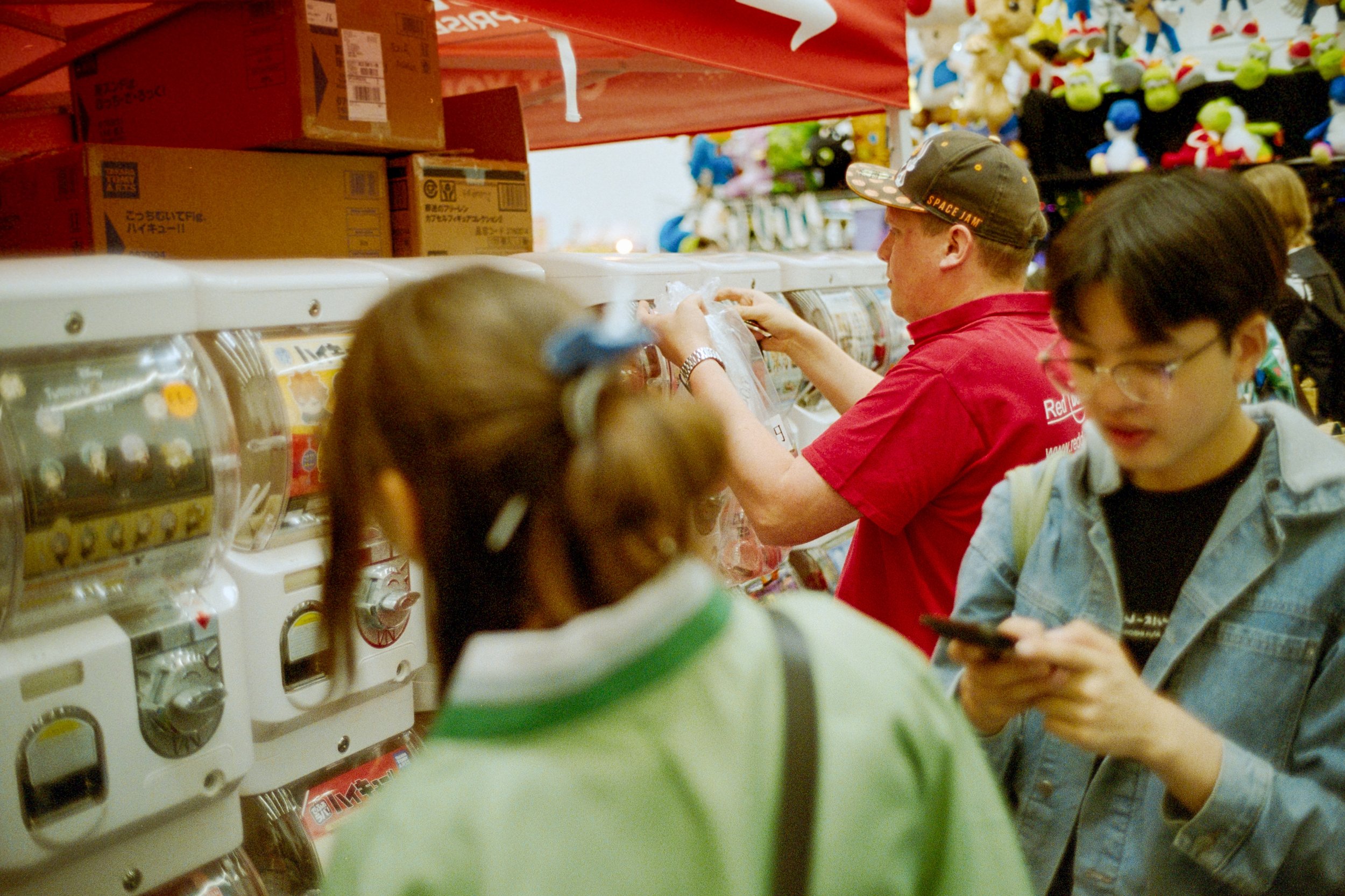 Menschen stehen an einer Gumball-Maschine in einem Spielzeugladen, einige schauen auf ihre Handys, andere suchen Preise.