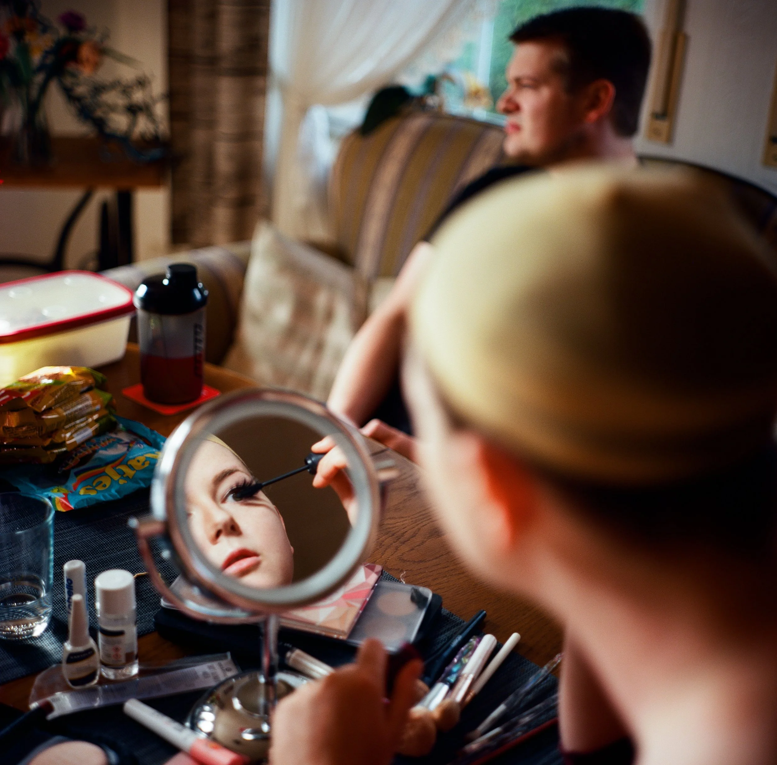 Eine Frau trägt Make-up auf ihr Auge auf, während sie in einem kleinen Spiegel schaut. Im Hintergrund sitzen ein Mann und eine andere Person am Tisch, während die Frau im Vordergrund beim Schminken beobachtet wird.