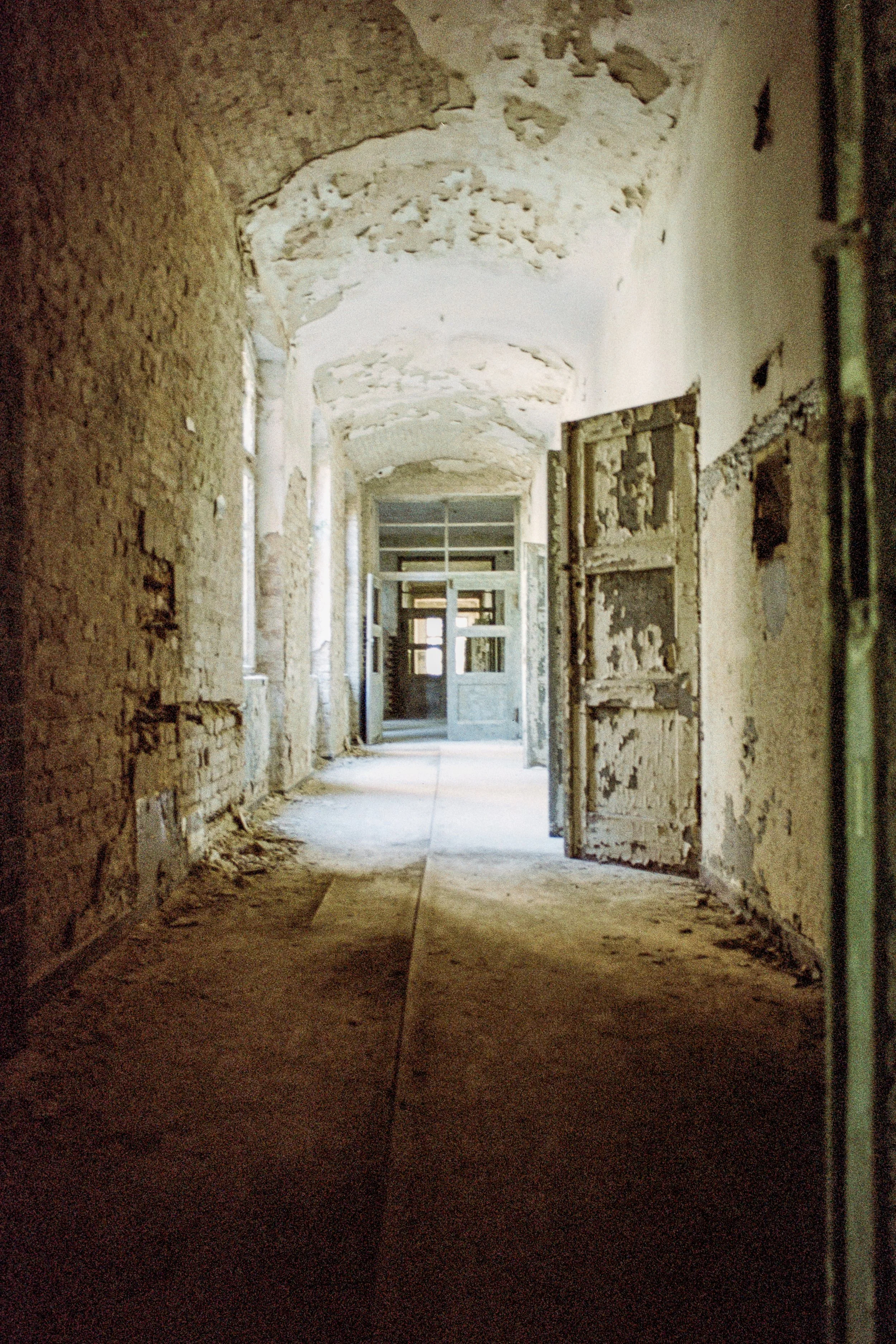 Ein verlassener, verfallener Flur in einem alten Gebäude, mit teilweise beschädigten Wänden und Türen, durch die Tageslicht einfällt.