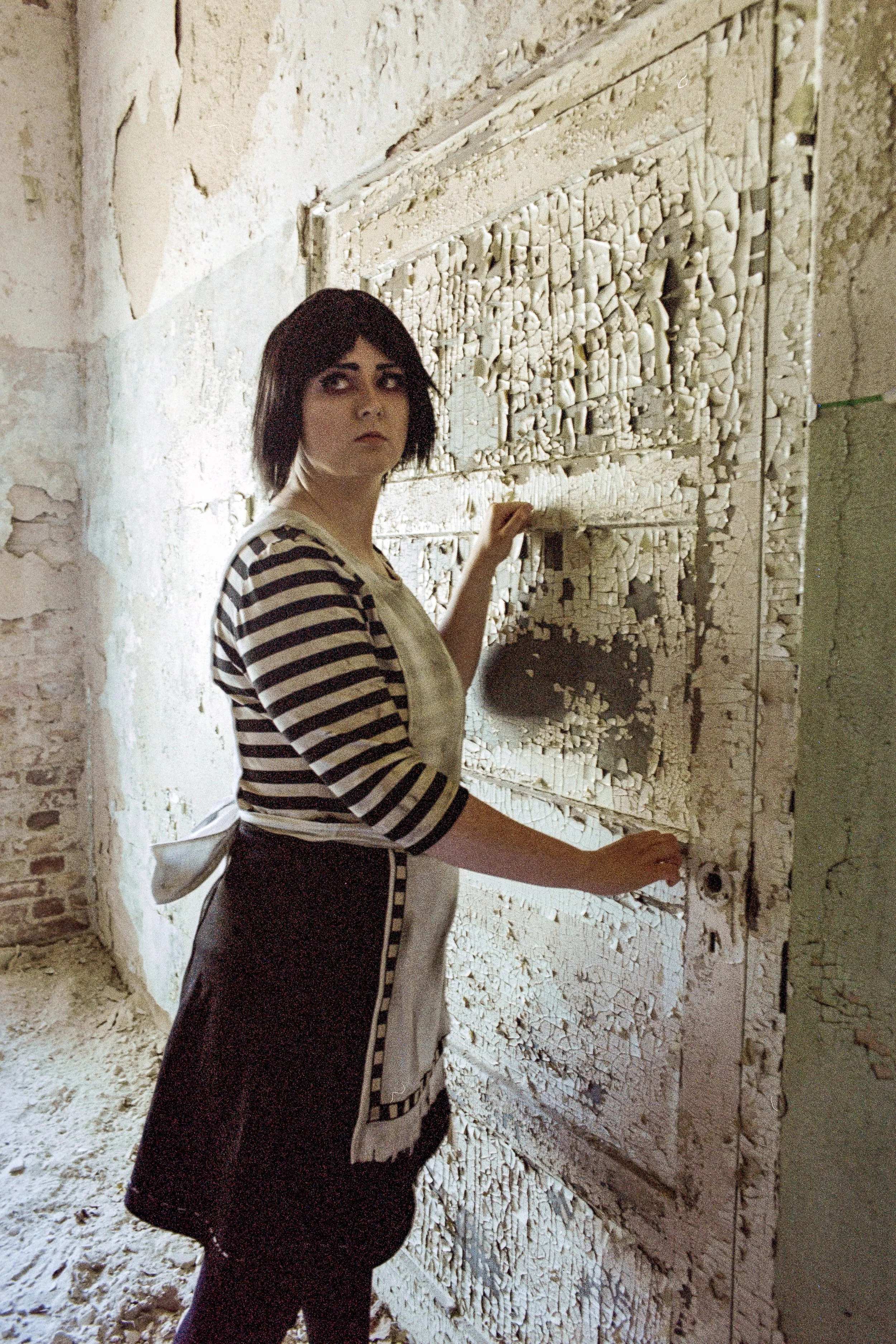 Eine Frau mit schwarzen Haaren, gestreiftem Oberteil und Schürze steht vor einer alten, zerfallenen Tür in einer verfallenen, ruinösen Umgebung.