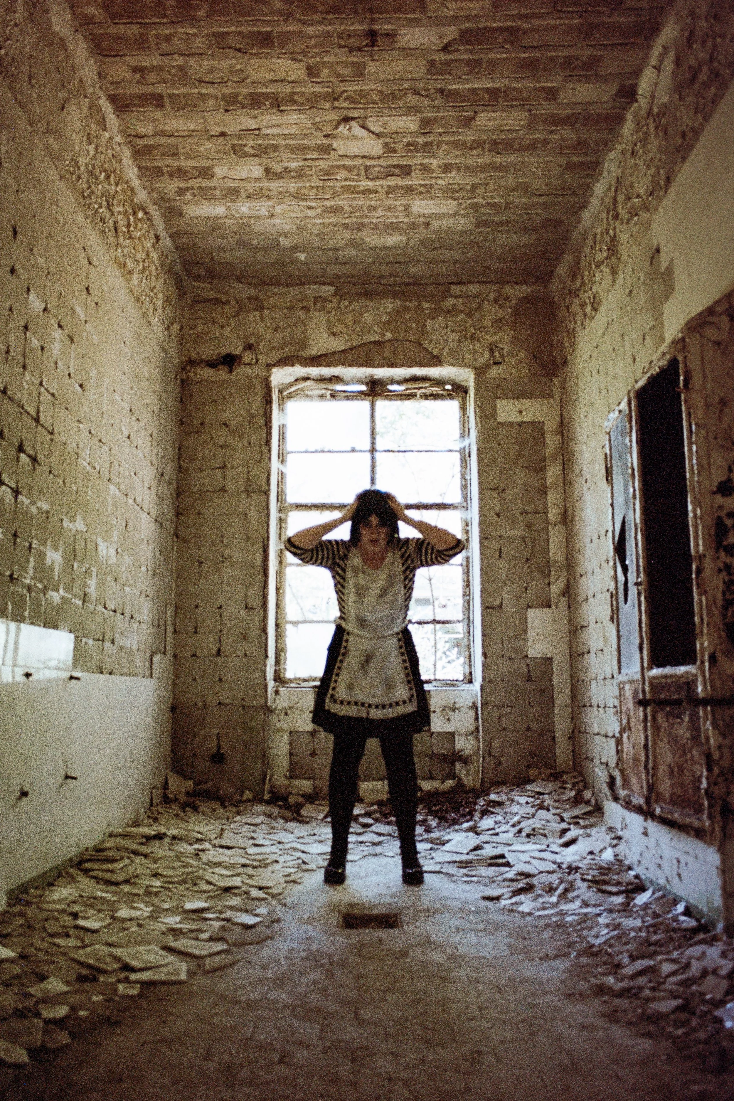 Kind in einem verfallenen, leeren Raum mit zerborstenen Wänden und Boden, vor einer Fensterfront, hält den Kopf mit beiden Händen, sozial, unheimlich, verlassen.