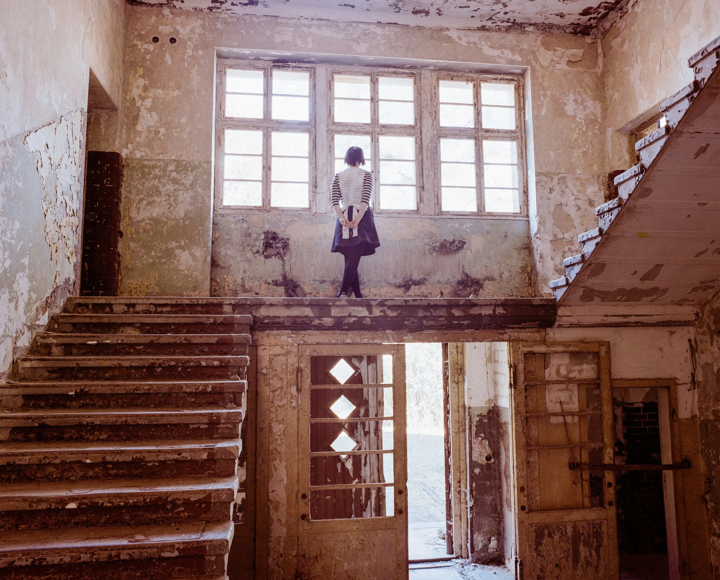 Eine junge Frau steht in einem verfallenen, alten Gebäude auf einer oberen Etage, vor einem großen Fenster, das Licht hereinlässt. Die Wände sind abgeblättert, und die Treppe ist abgenutzt.
