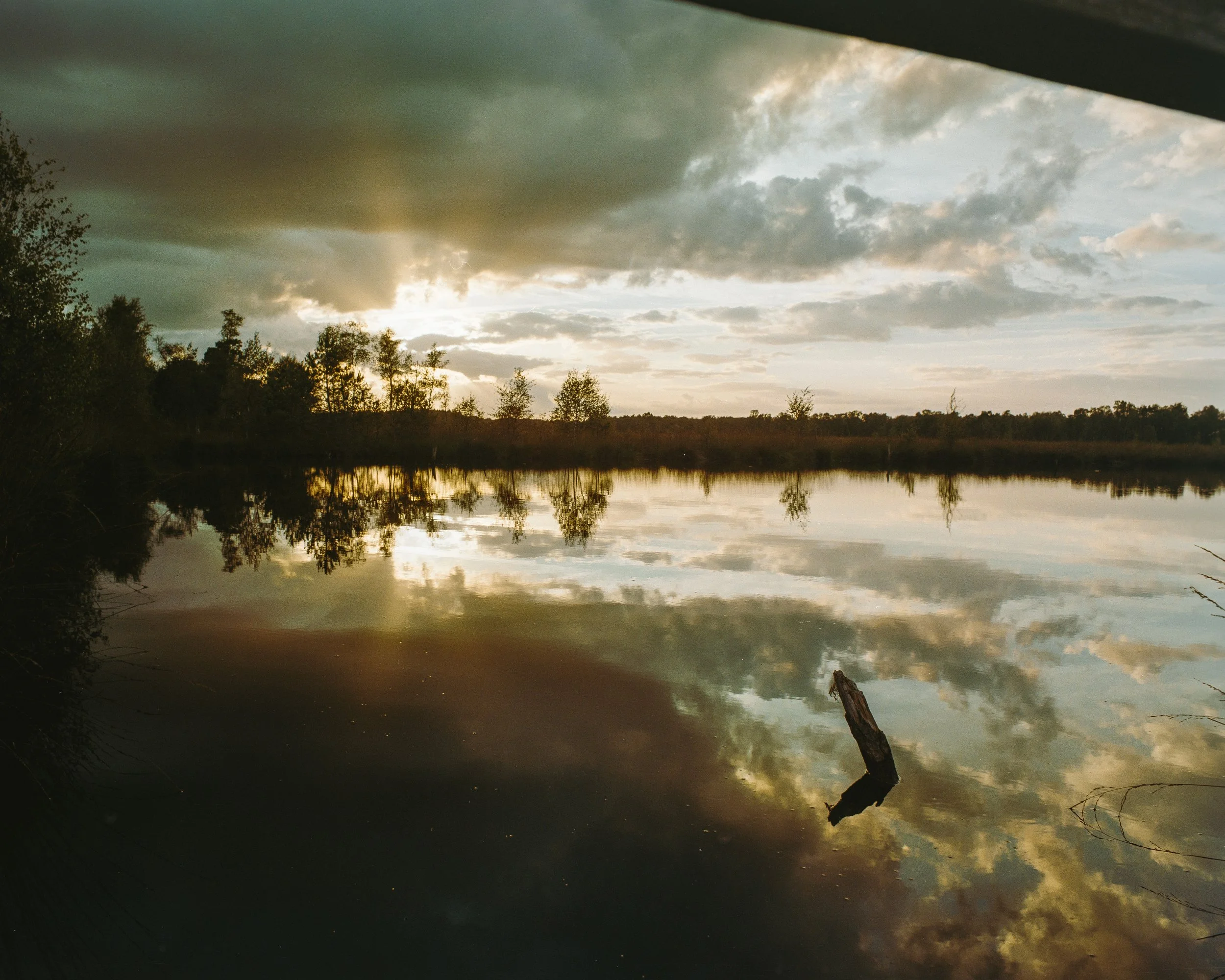 Ein ruhiger See bei Sonnenuntergang mit Bäumen am Horizont und spiegelnder Wasseroberfläche, umgeben von dunklen Wolken.