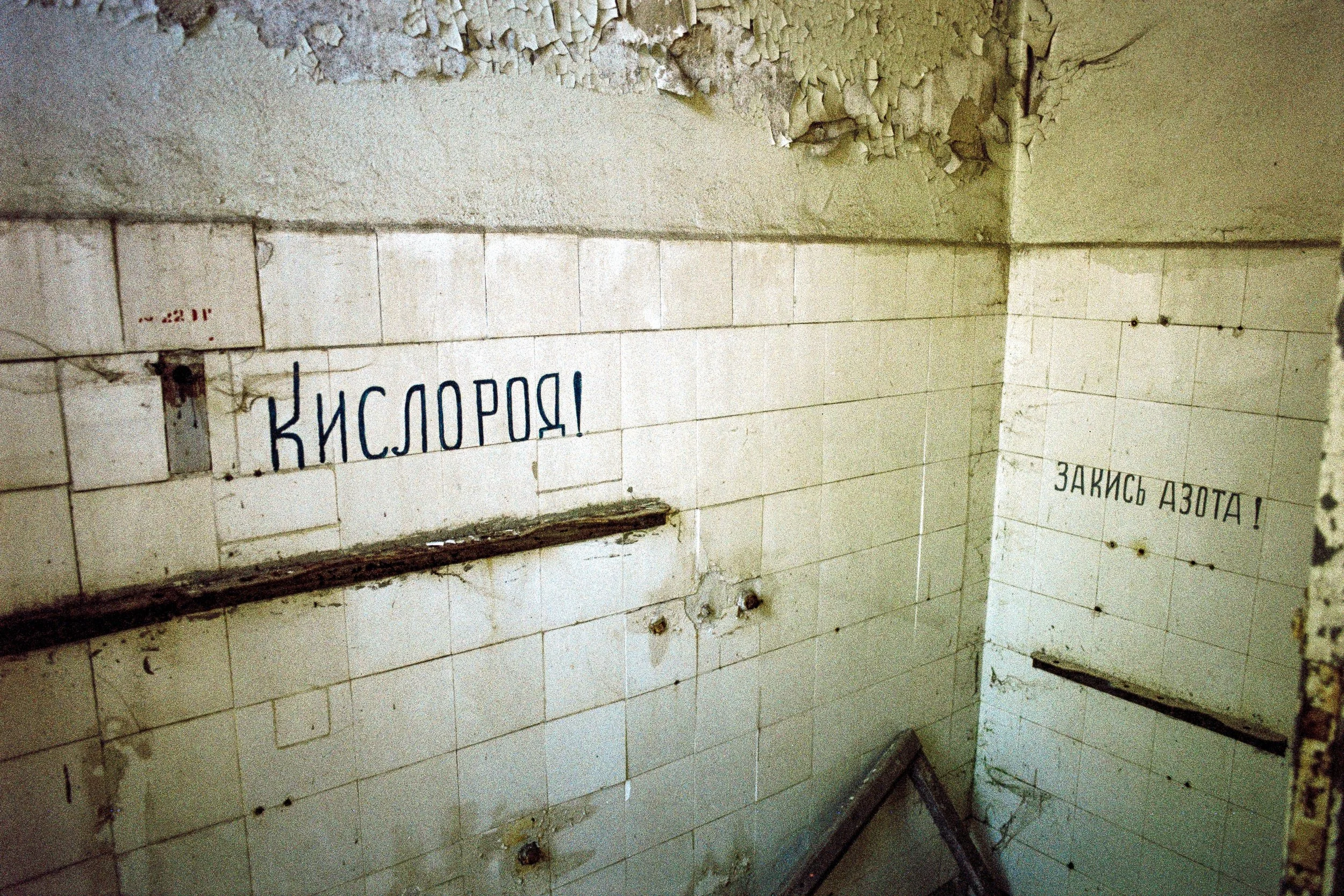 Verwüstete, alte Badezimmerwand mit Fliesen und Graffiti mit den Worten „КИСЛОРОД!“ und „ЗАКИСЬ АЗОТА!“, obscur und verfallen.