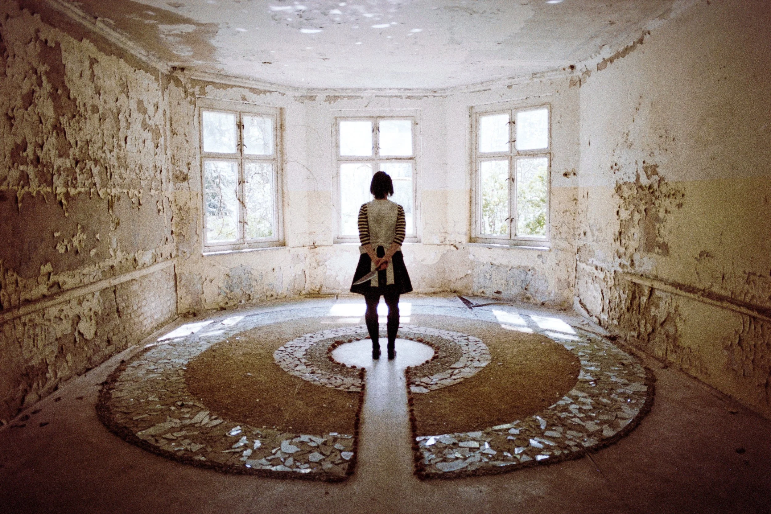 Ein Mensch steht in einem verlassenen, alten Raum mit zerbröckelnden Wänden und vier großen Fenstern, der Raum hat einen kunstvollen Mosaikboden, der von Sonnenlicht durch die Fenster beleuchtet wird.