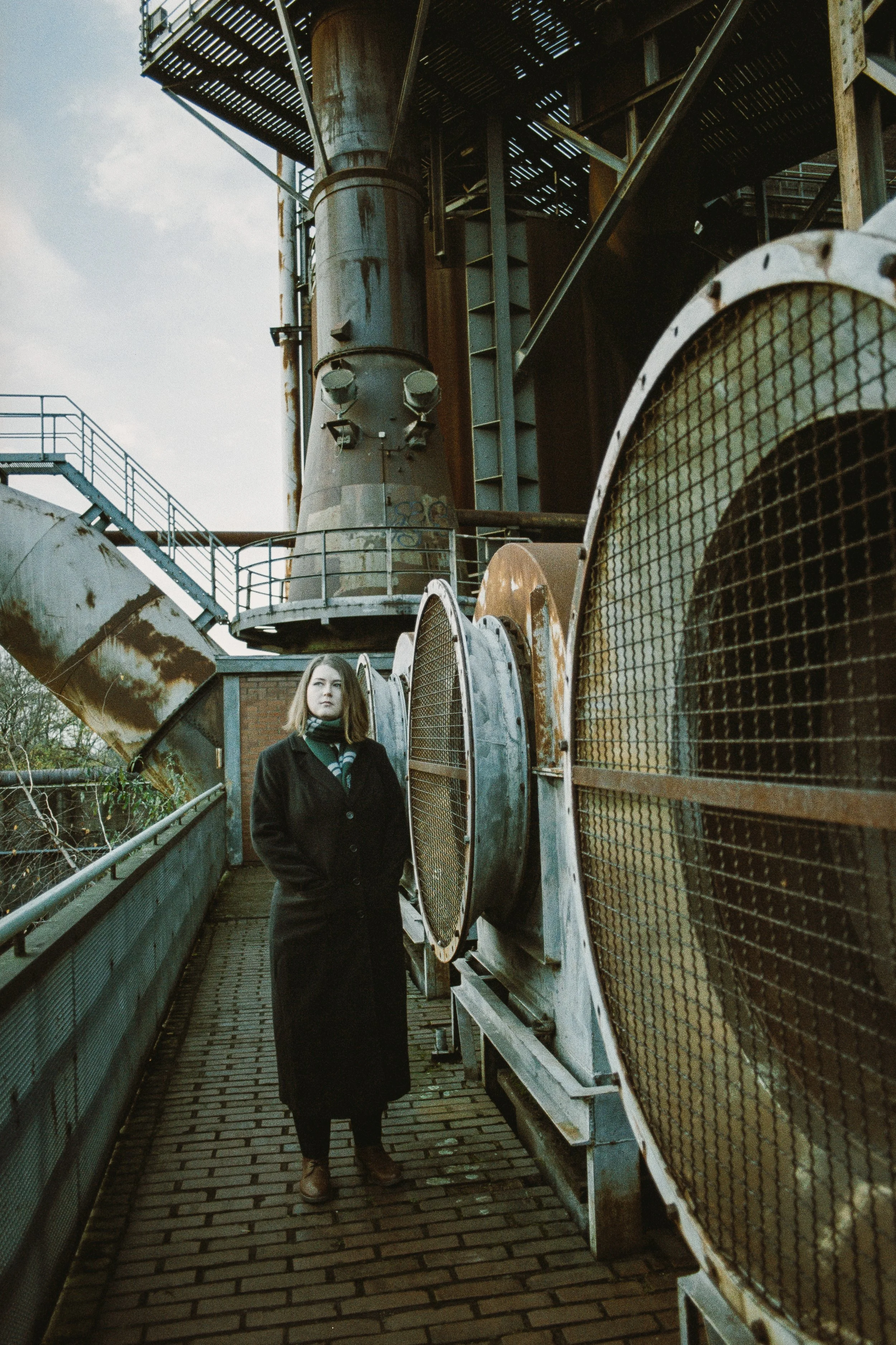 Eine Frau steht auf einer Brücke neben großen, verrosteten industriellen Lüftungsöffnungen bei einem alten, rustikalen Fabrikgebäude.