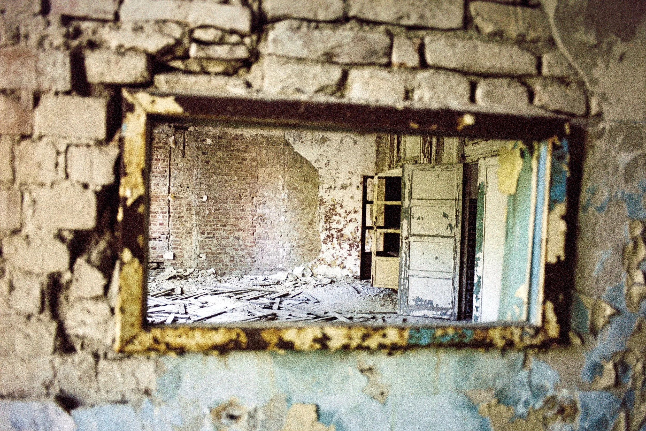 Blick durch ein kleines, verrostetes Fenster in eine verlassene, verfallene Raum mit abblätternder Farbe und zerbrochenen Böden.