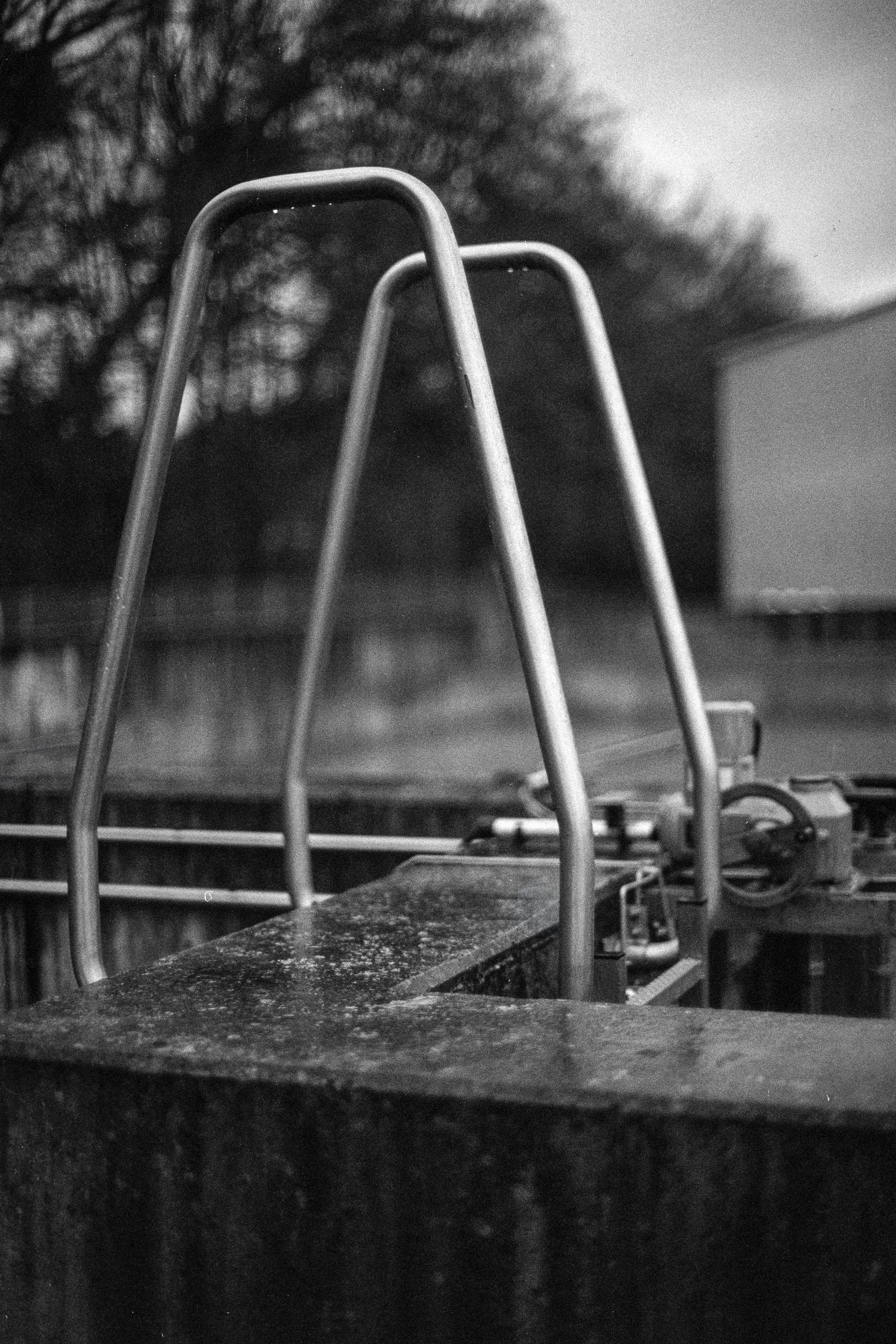 Ein schwarzer und weißer Schwarzweiß-Foto eines metallenen Schwimmbad-Standrings, der auf einem Wasserbecken steht, im Hintergrund sind Bäume und ein Gebäude sichtbar.