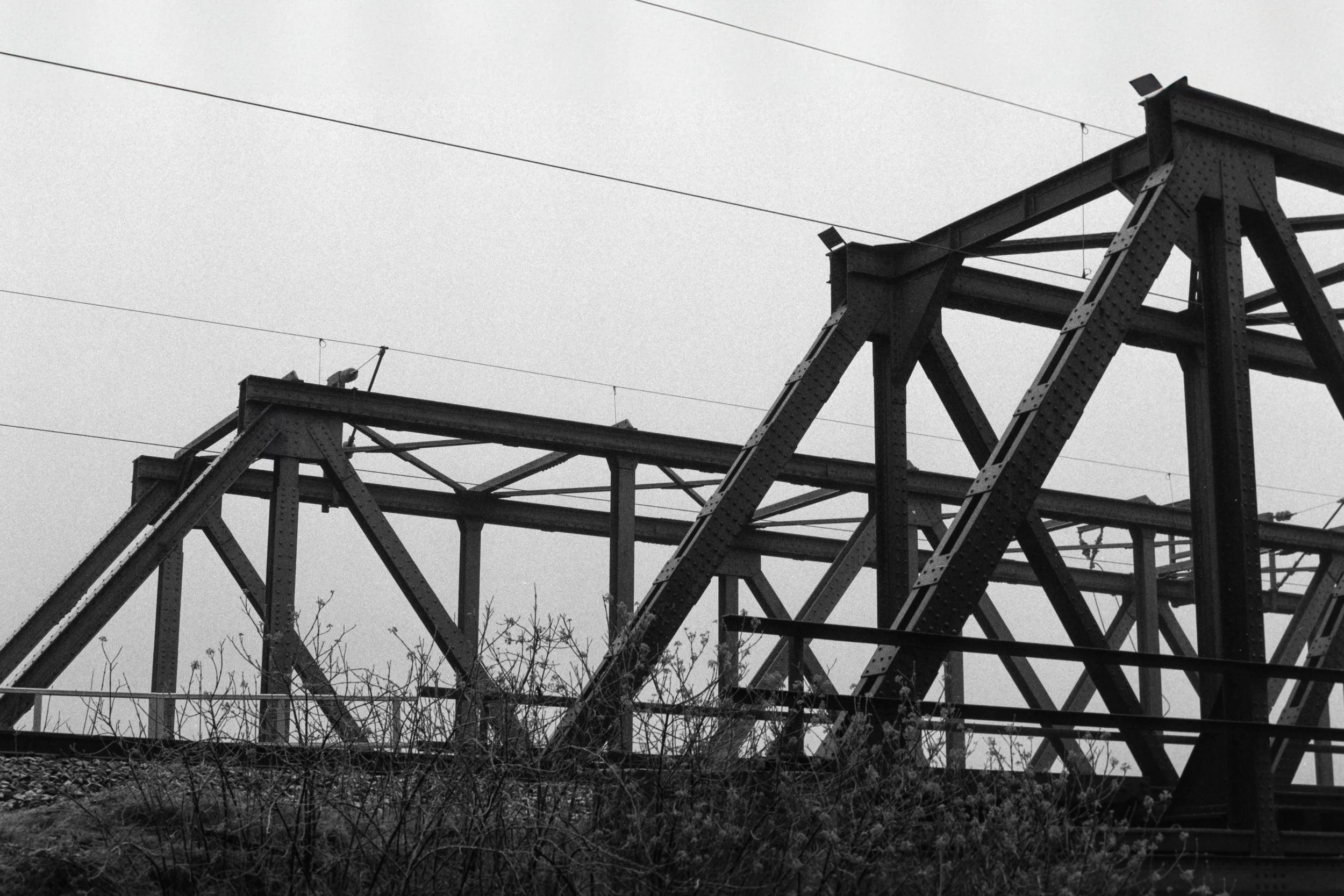 Schwarz-weiß-Foto einer alten Eisenbahnbrücke mit Metallträgern und Kabeln gegen einen grauen Himmel.