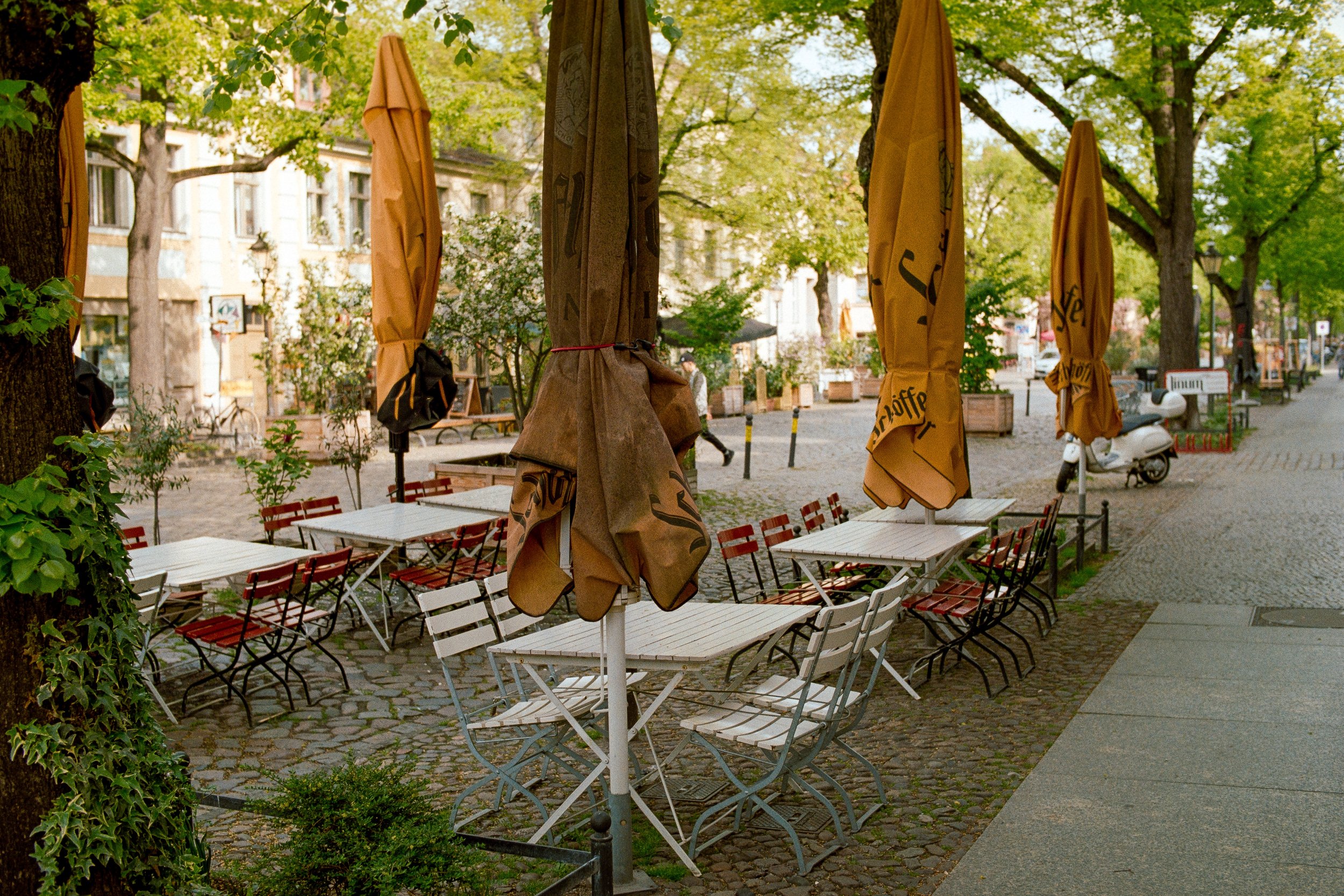Leere Straßencafétische mit umgekrempelten Sonnenschirmen und verschobenen Stühlen, umgeben von Bäumen und Gebäuden in einer ruhigen Stadtstraße.