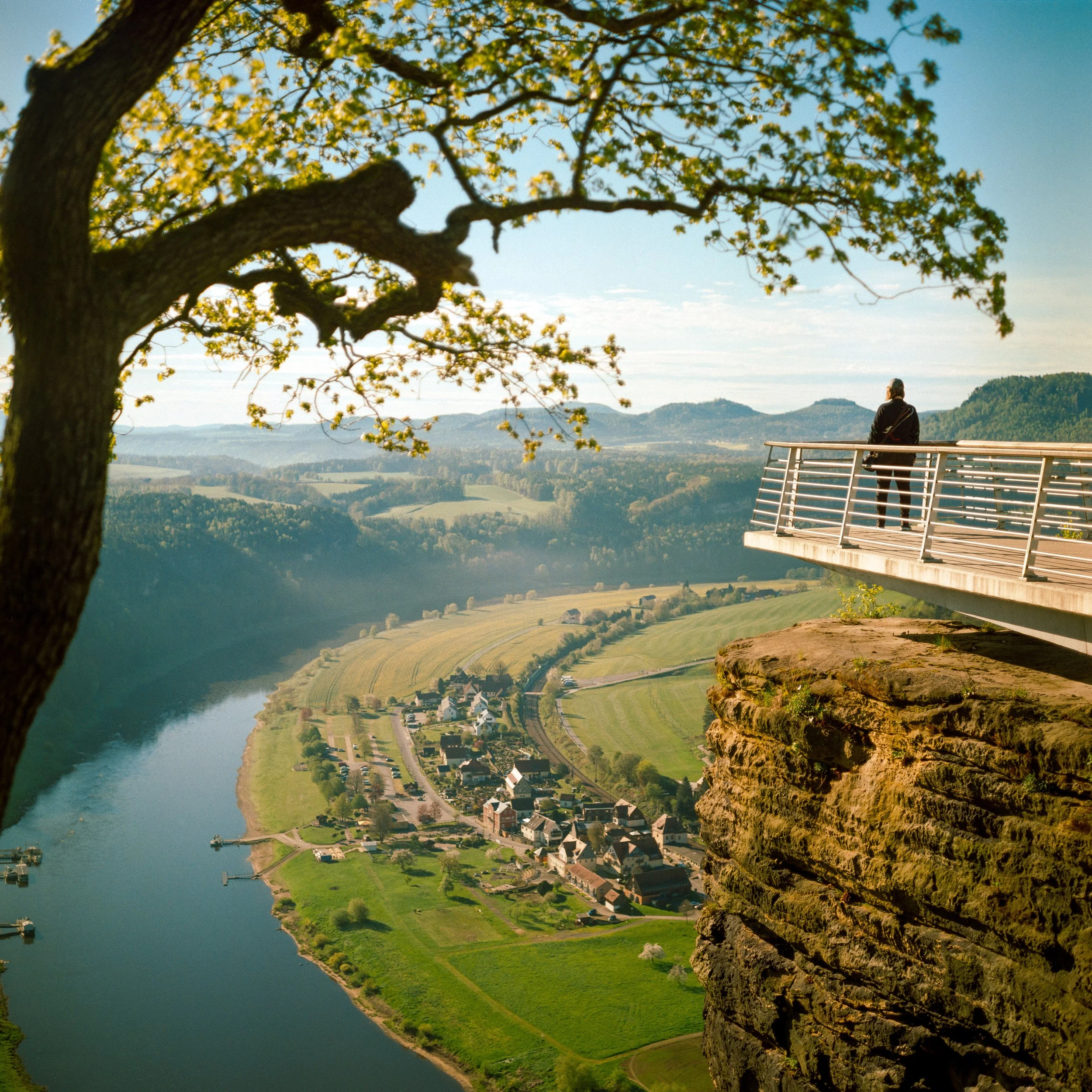 Person auf einer Aussichtsplattform über einem Fluss und einer kleinen Stadt in einer grünen, hügeligen Landschaft, mit Baum im Vordergrund.