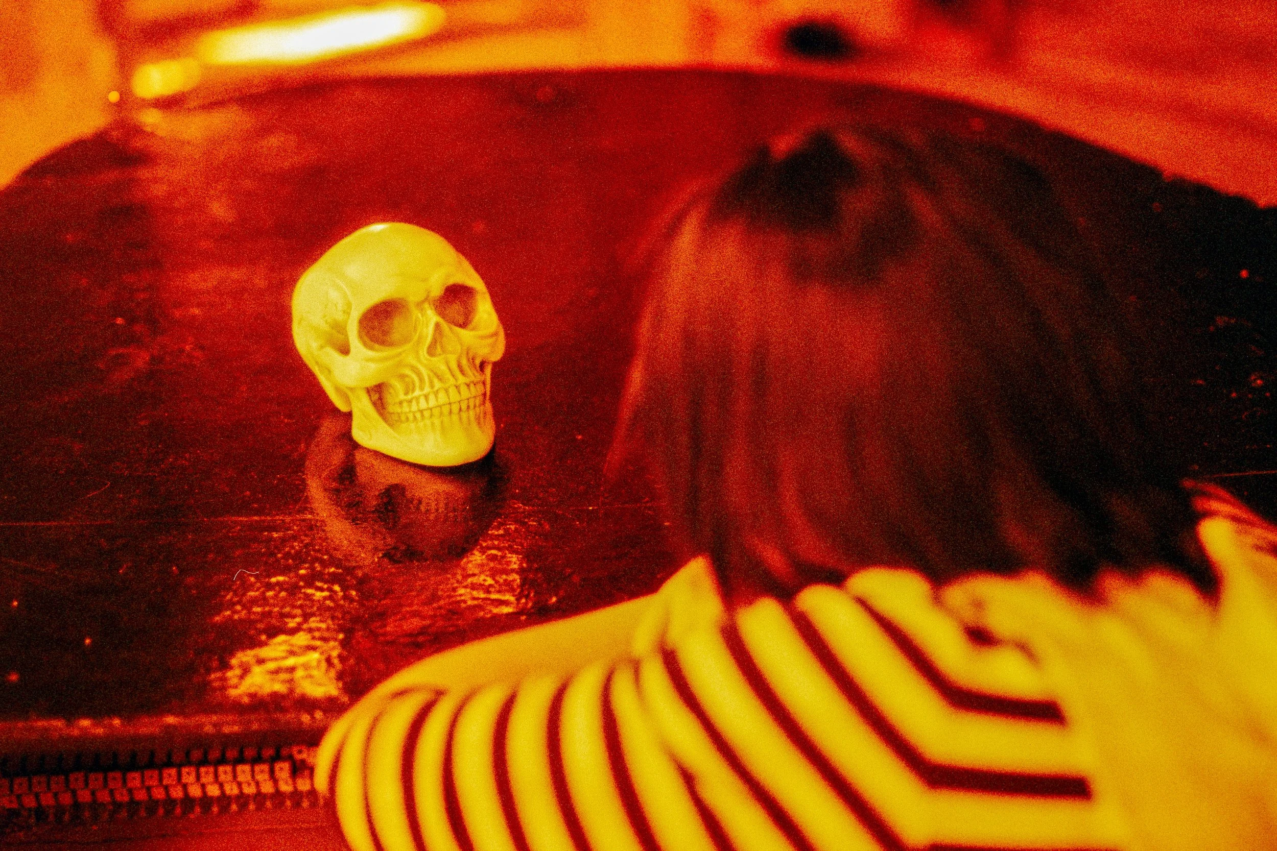 Eine Person mit braunen Haaren, die eine schwarze und weiße gestreifte Jacke trägt, blickt auf eine gelbe Schädel-Statue, die in einem roten Licht auf einem dunklen Tisch liegt.