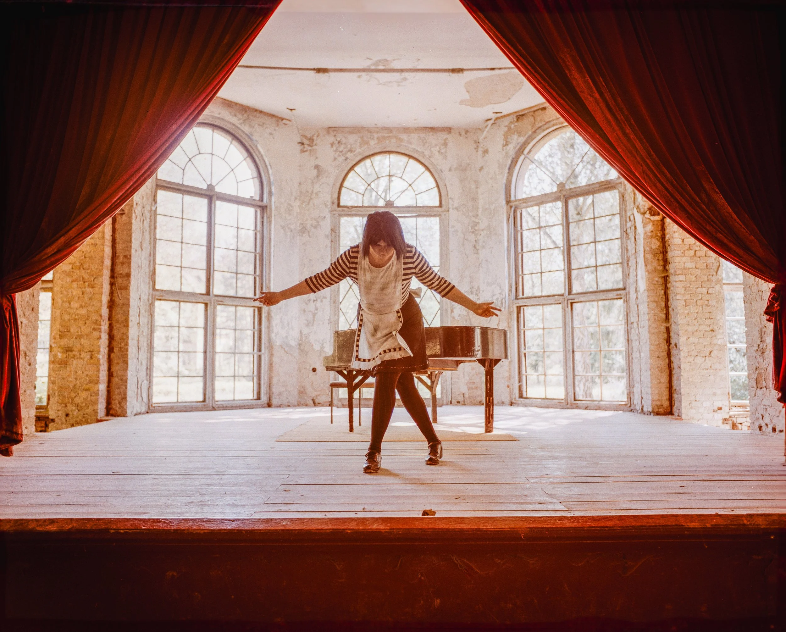 Eine Person steht auf einer alten Bühne mit roten Vorhängen und großen Fenstern im Hintergrund, die ein altes, verwittertes Gebäude zeigen.