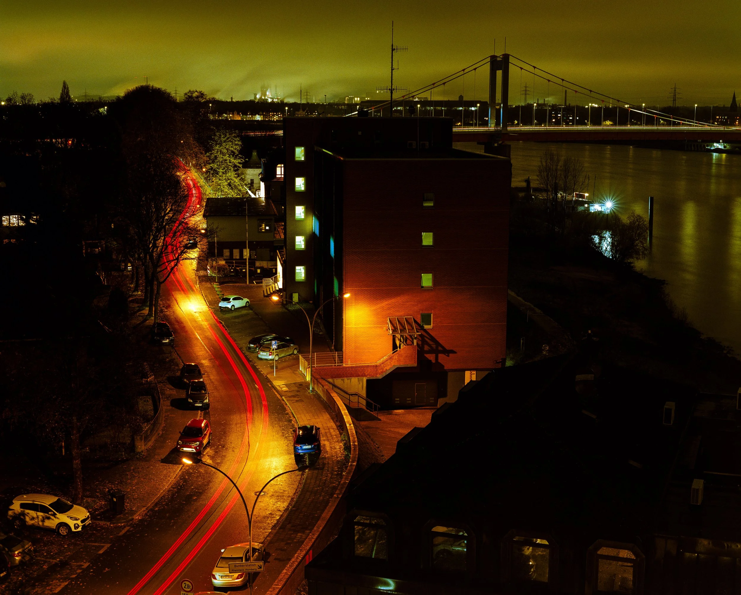 Nachtaufnahme einer Stadtansicht mit Gebäuden, Brücke über einen Fluss, beleuchteter Straße mit Fahrzeugen, und einer bewölkten grünlich leuchtenden Himmel.