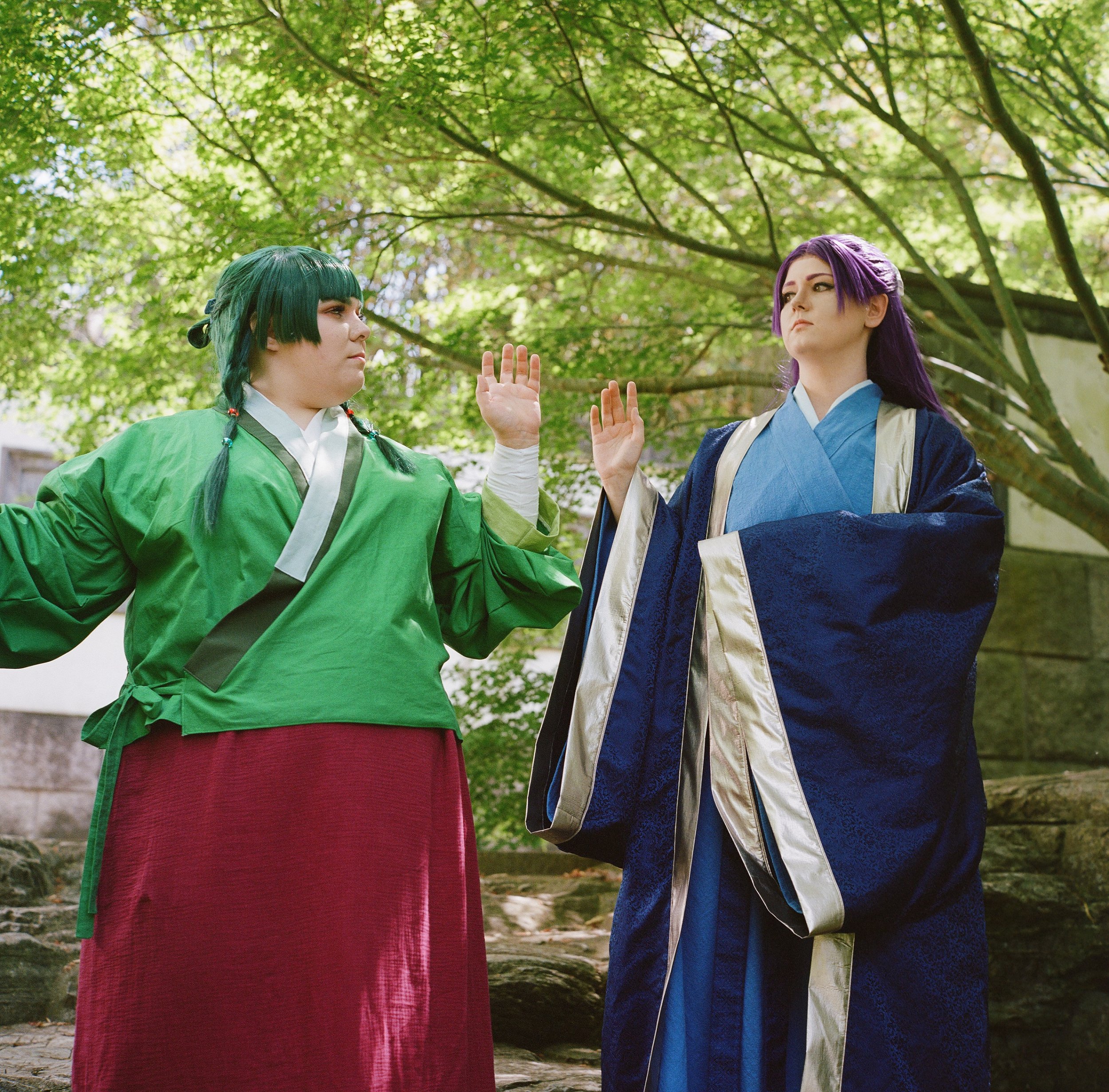 Zwei Menschen in traditionellen japanischen Kostümen stehen sich gegenüber und berühren sich mit den Handflächen, im Freien unter einem Baum mit grünen Blättern.