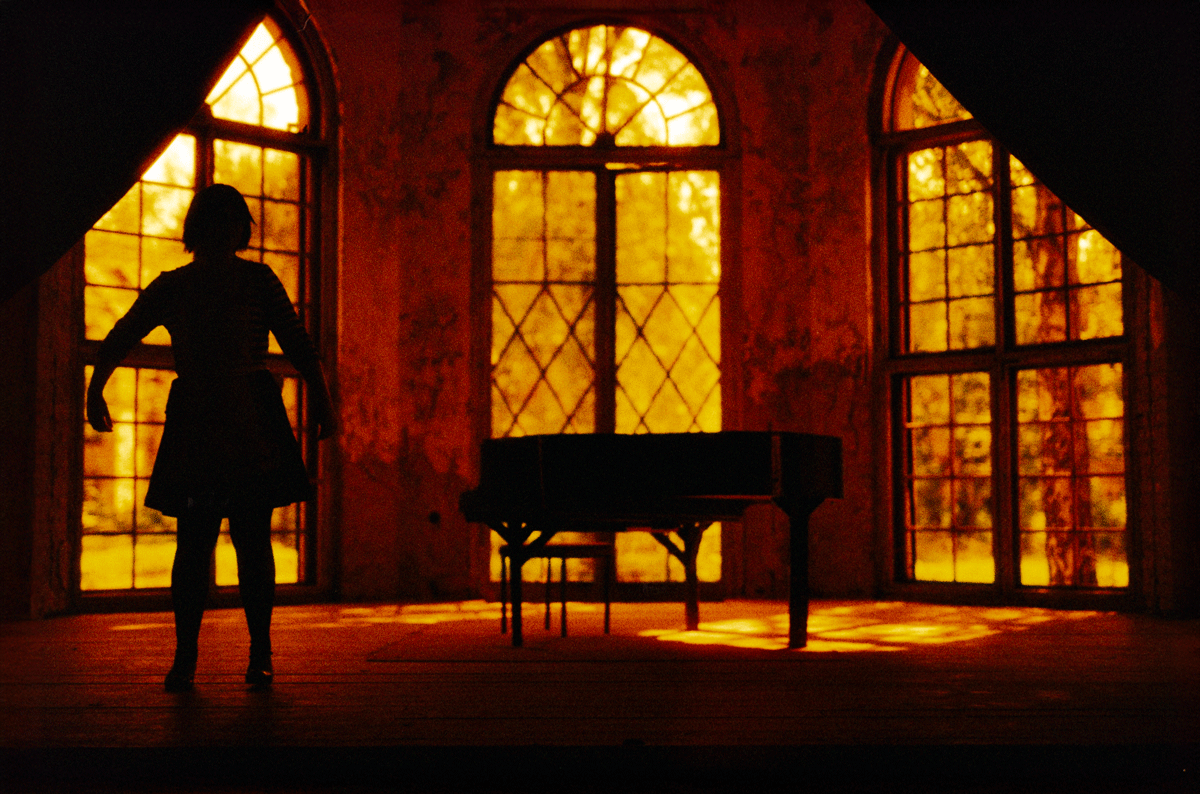 Ein dunkler Raum mit großen fenstern und einer Silhouette einer Person, daneben ein Klavier.