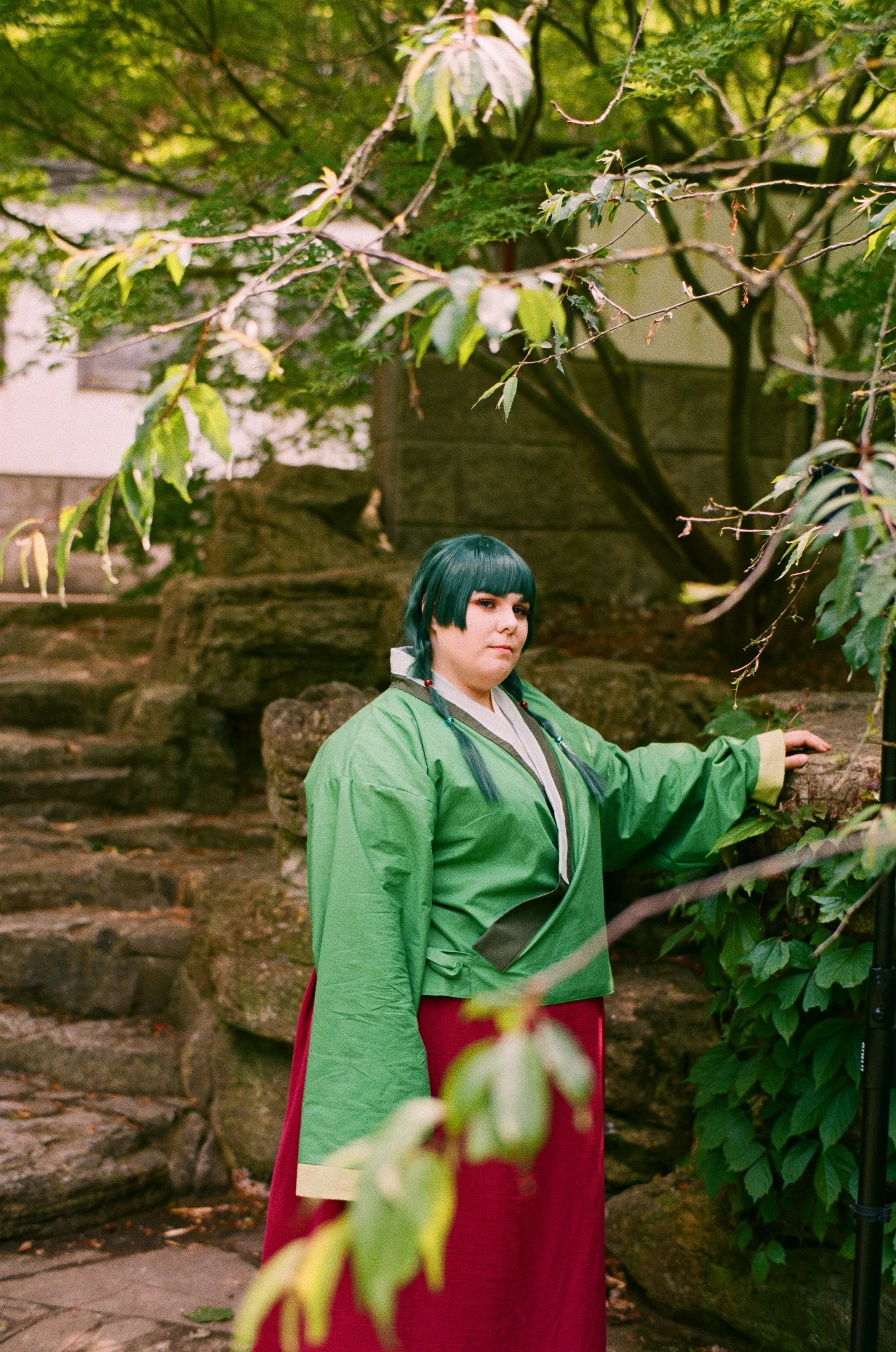 Person in Cosplay-Kleidung mit grüner Jacke und roten Rock vor natürlichen Hintergrund mit Steinen und Bäumen.