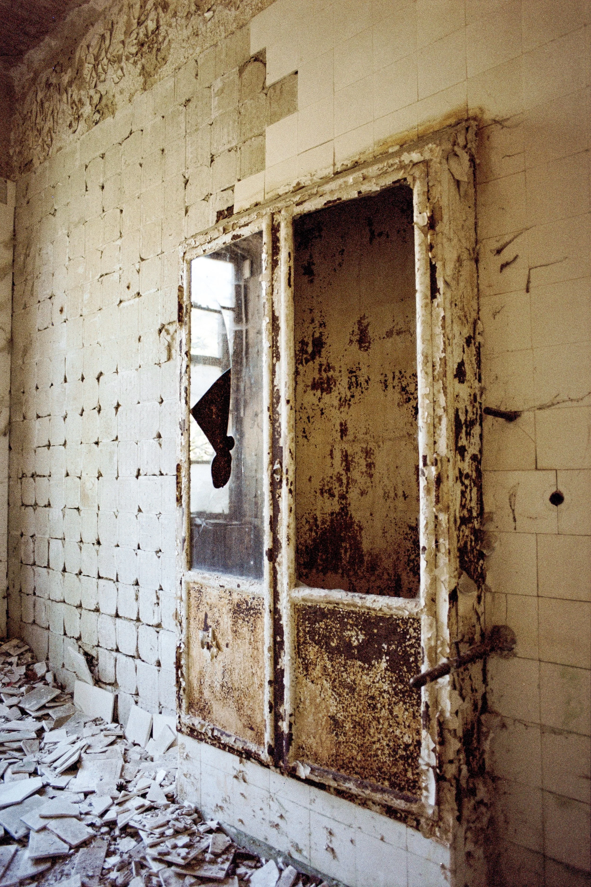 Verrosteter, alter Aufzugsschacht in einer verfallenen, staubigen Gebäudewand, durch das Scheiben Glas ist eine schwarze Hand zu sehen, umgeben von zerstörtem Mauerwerk und Trümmern auf dem Boden.