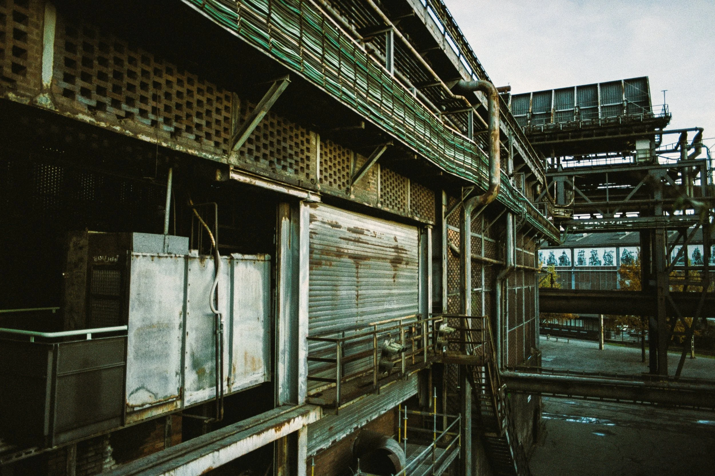 Verfallene Industrieanlage mit rostigen Metallstrukturen, Rohrleitungen und verschlossenen Türen, im Freien bei bewölktem Himmel.