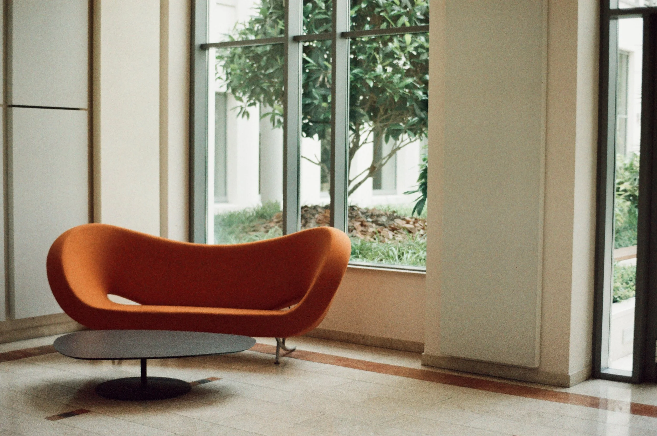 Ein modernes, orangefarbenes Sofa vor großen Fenstern mit Blick auf einen Garten mit Sträuchern und Bäumen.