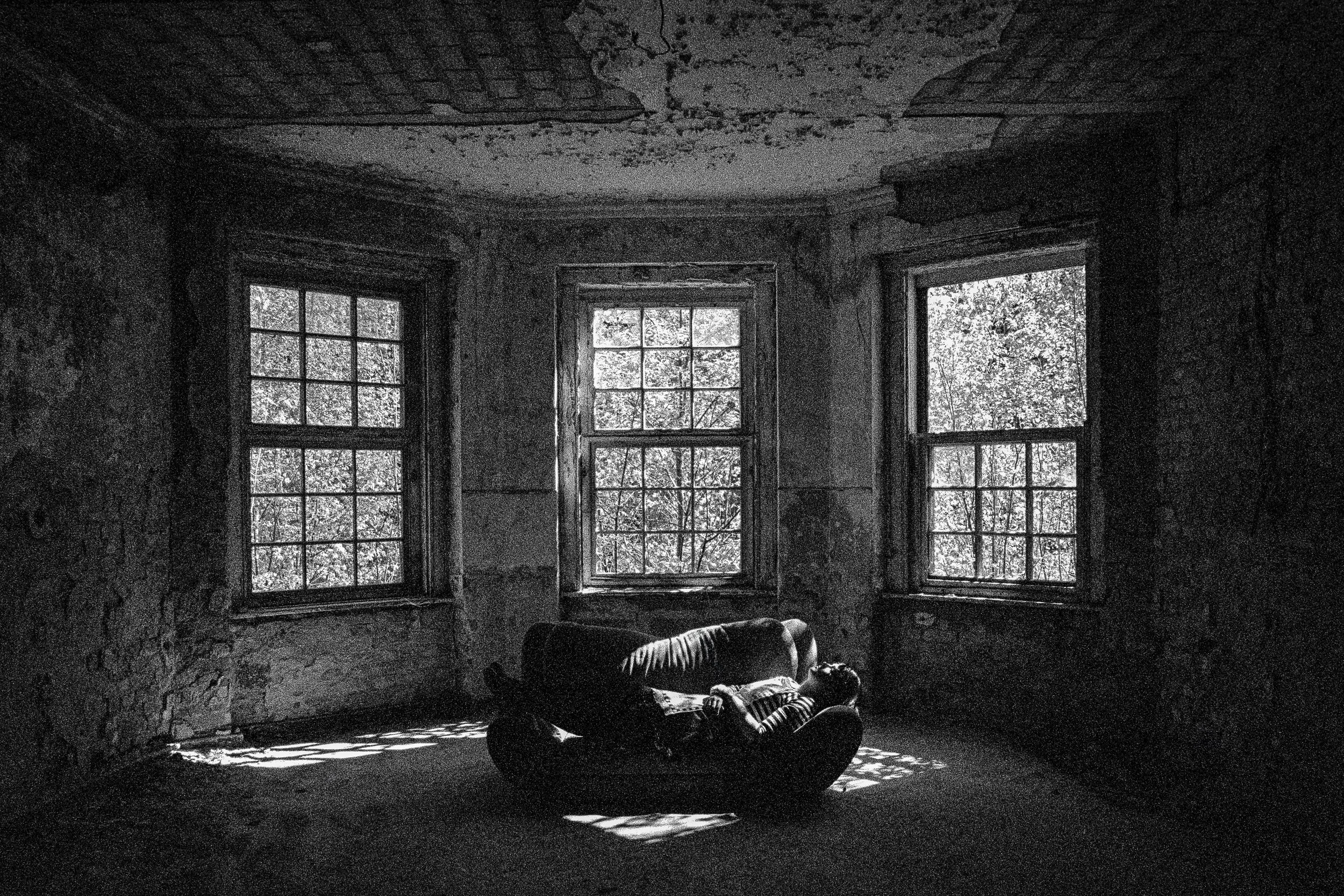 Schwarz-weiß-Bild eines alten, verlassenen Zimmers mit drei Fenstern, durch die Licht und Bäume sichtbar sind. Auf einer Couch liegt eine Person, die sich ausruht.