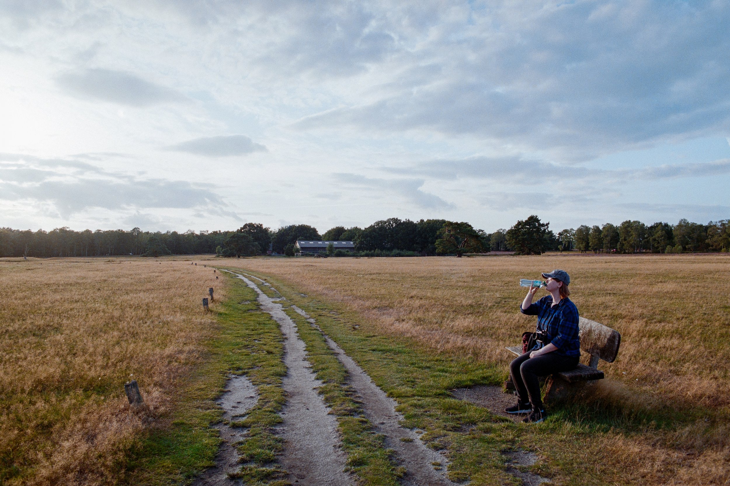 Frau sitzt auf einer Bank in einer weiten Wiesenlandschaft und trinkt Wasser.
