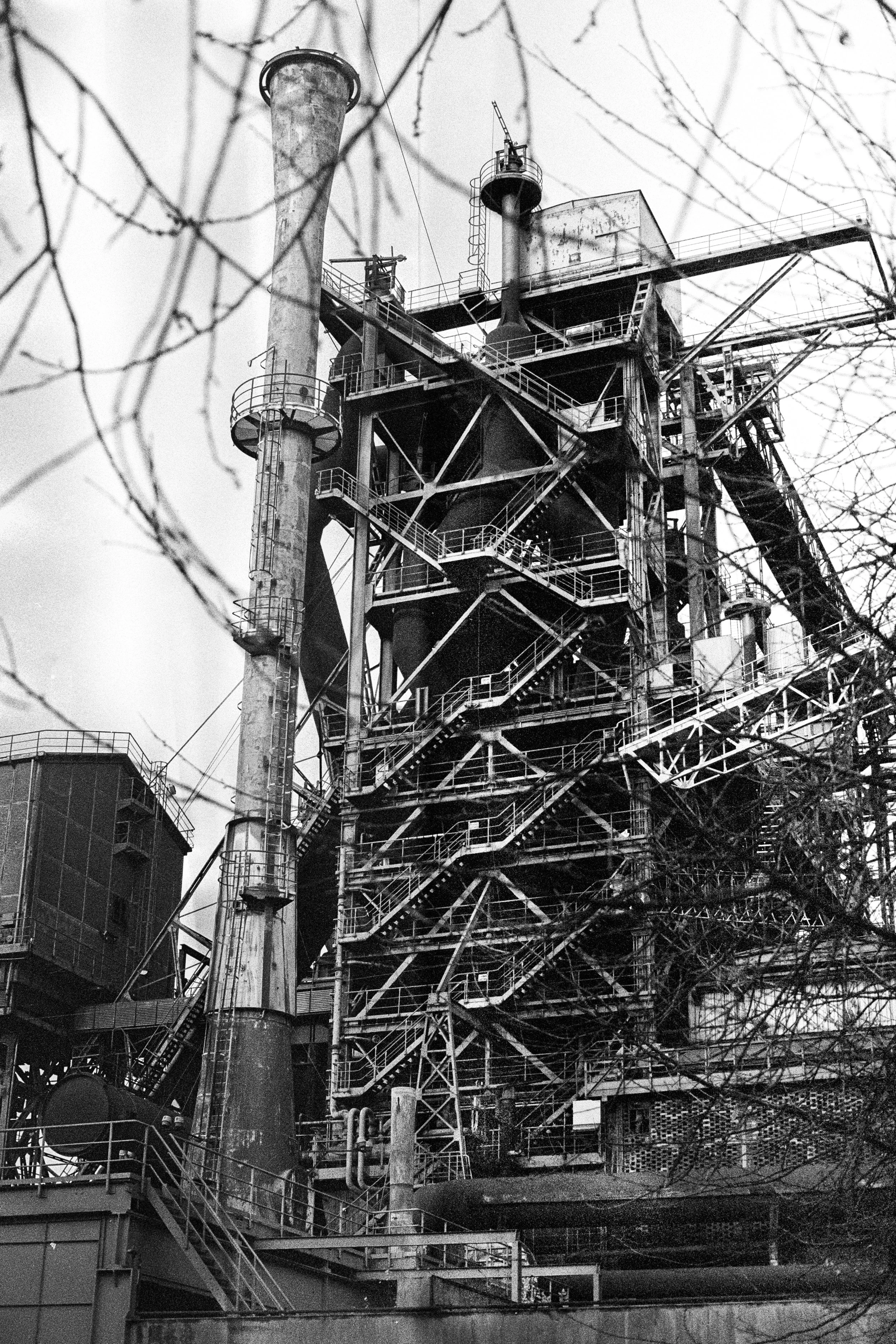 Schwarz-Weiß-Foto eines alten Industrieofens oder Kraftwerks mit mehreren Rohren und Stufen, umgeben von dünnen Zweigen.