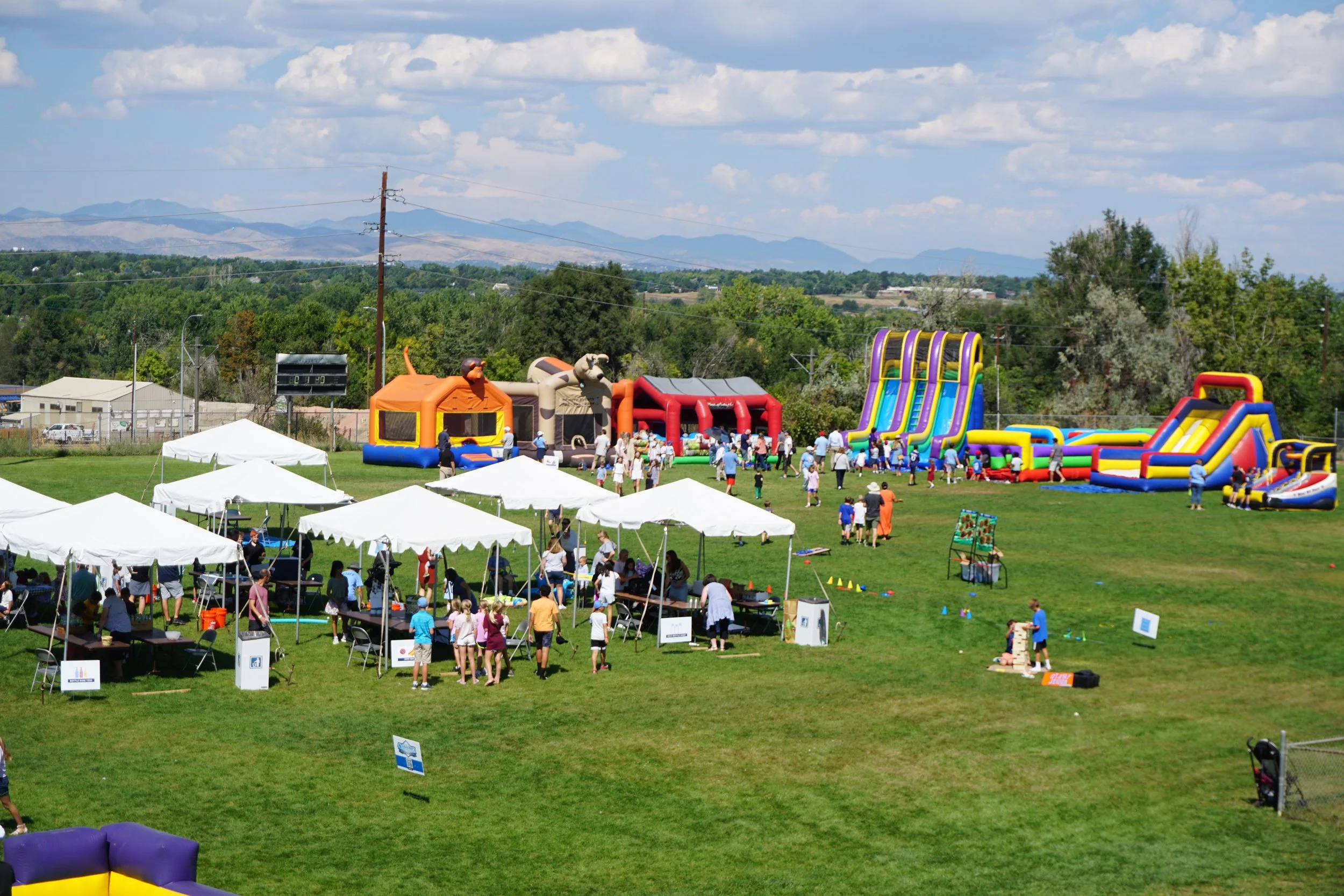 A large outdoor event featuring multiple inflatable bounce houses, slides, and tents with many attendees, children, and adults on a grassy field with trees and mountains in the background under a partly cloudy sky.