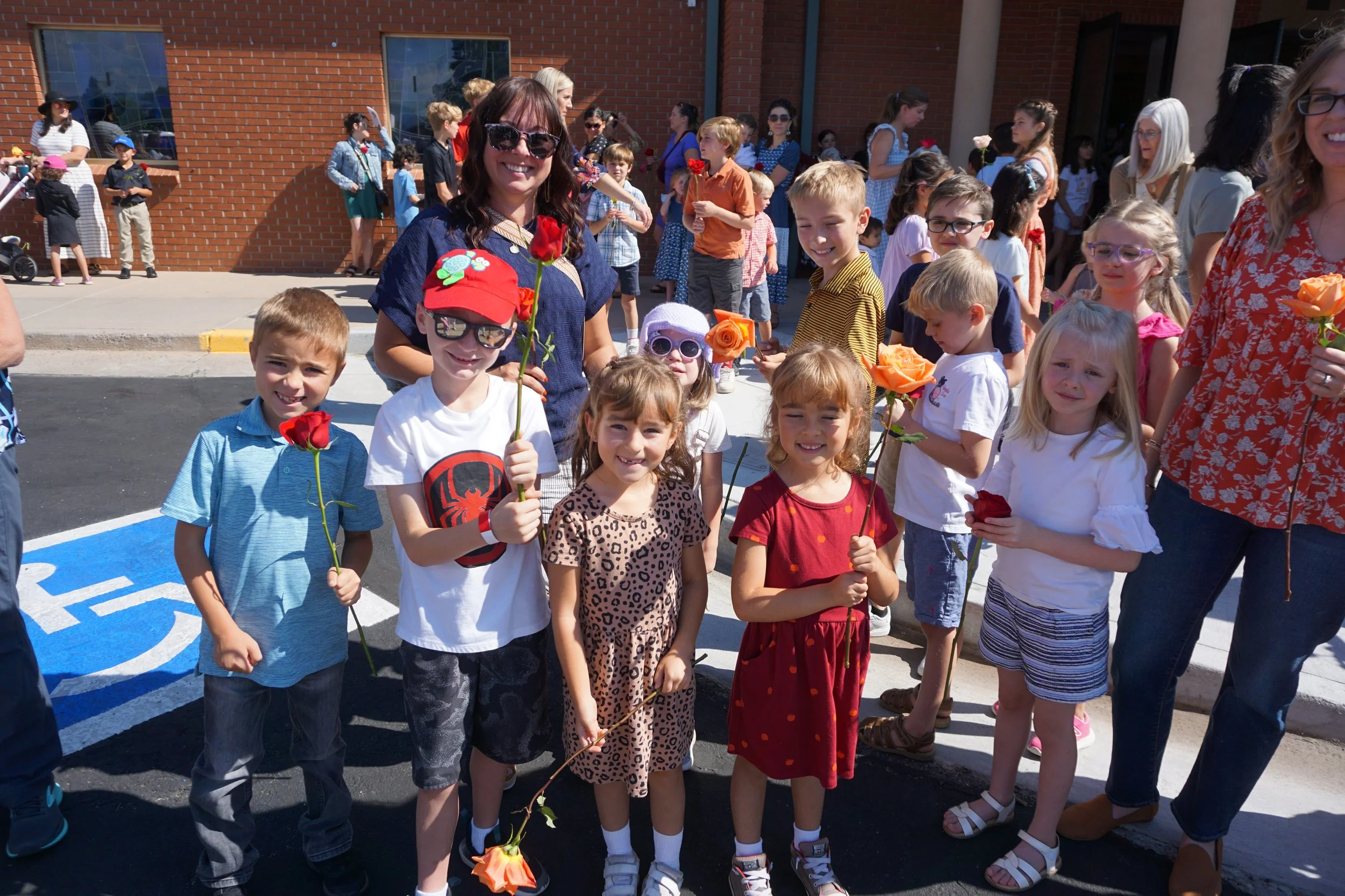 A group of children and adults gathered outside a school building, holding roses and flowers during a celebration or event.