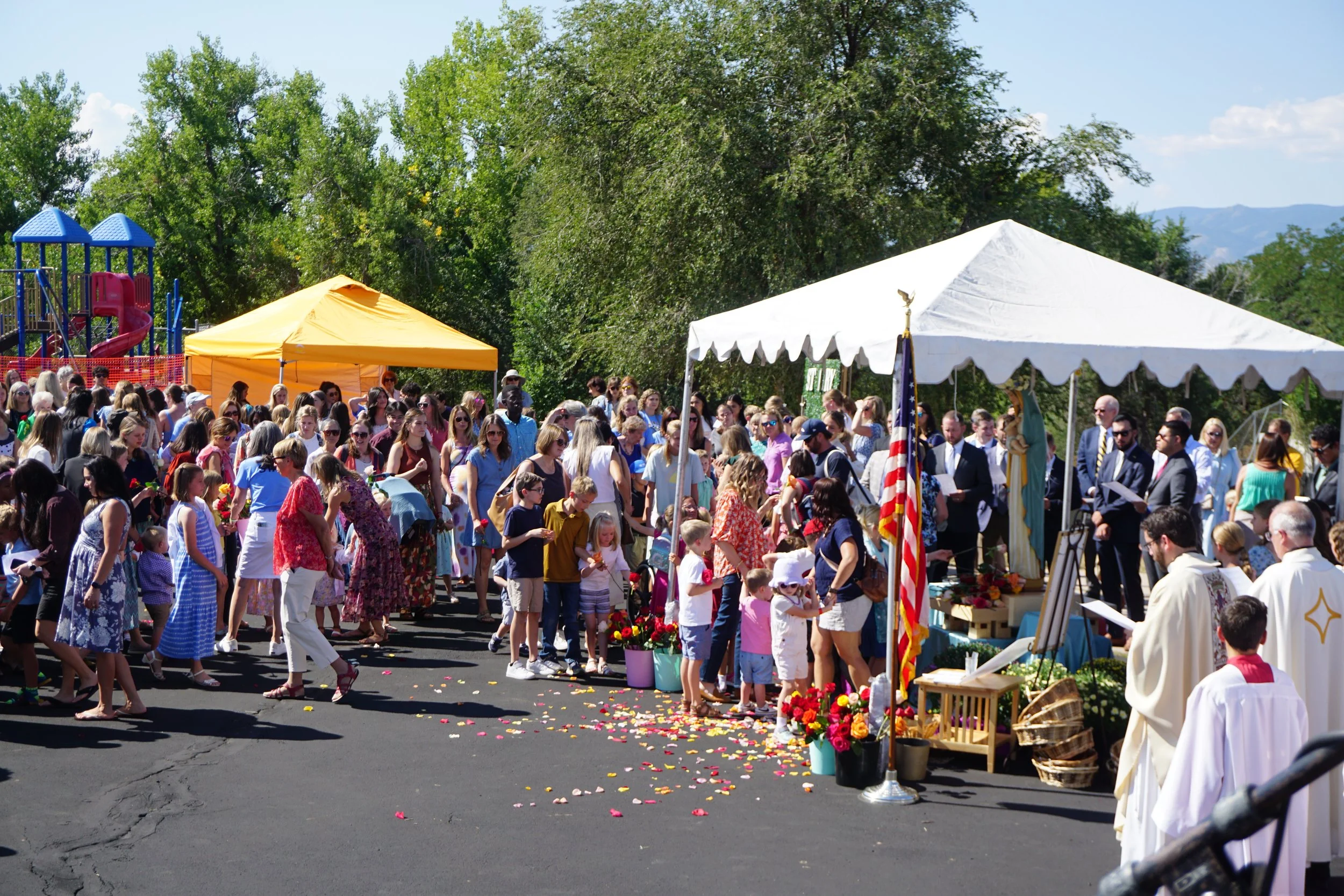 Crowd of people gathered outdoors at a religious or memorial event, with colorful flowers and flags, tents, and a playground in the background on a sunny day.