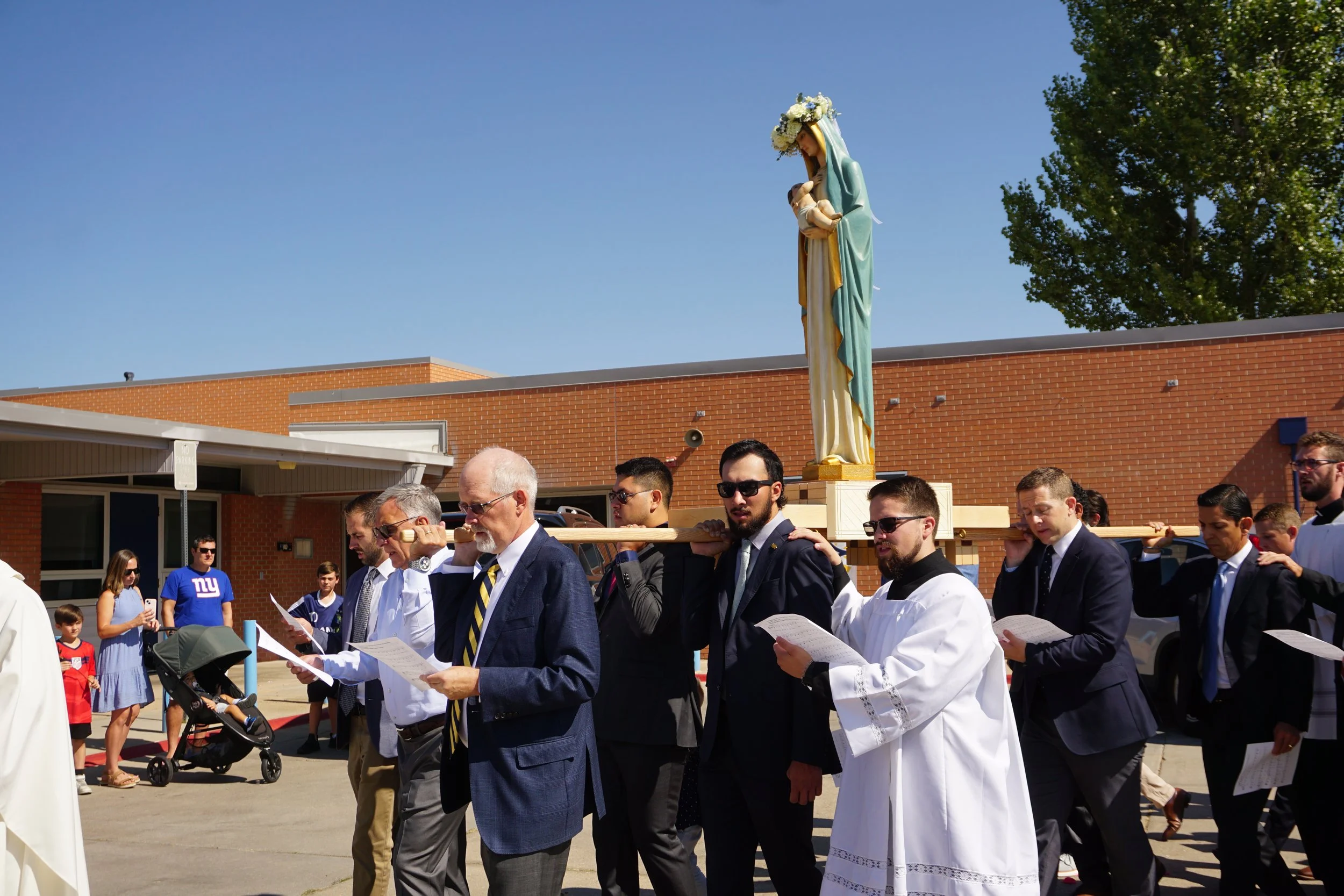 People participating in a religious procession, carrying a statue of the Virgin Mary with baby Jesus during an outdoor event on a bright sunny day. The participants are dressed in formal attire, and there are spectators and children in the background