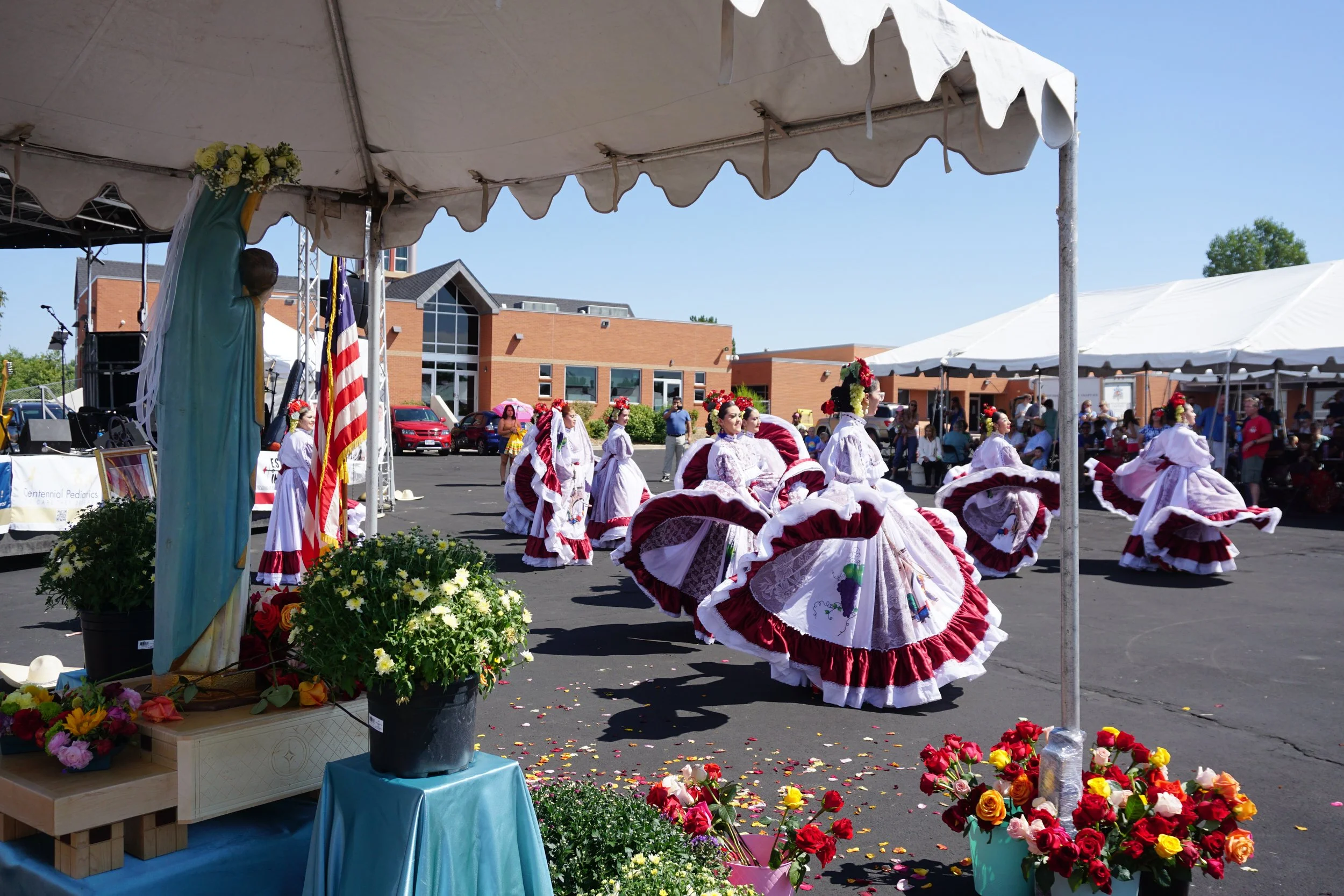 Group of women dressed in traditional Spanish Flamenco dresses performing a dance at an outdoor event, decorated with flowers and banners.