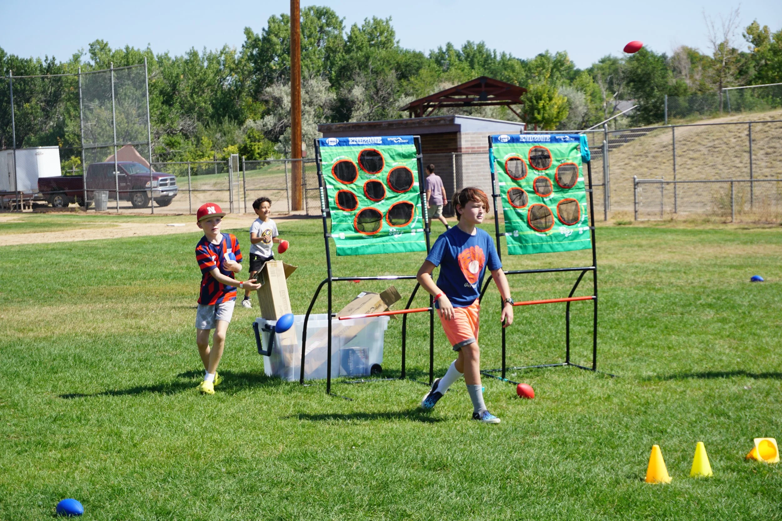 Children playing a dart-throwing game outdoors on a grassy field, with obstacles and cones set up for a game, and trees in the background.