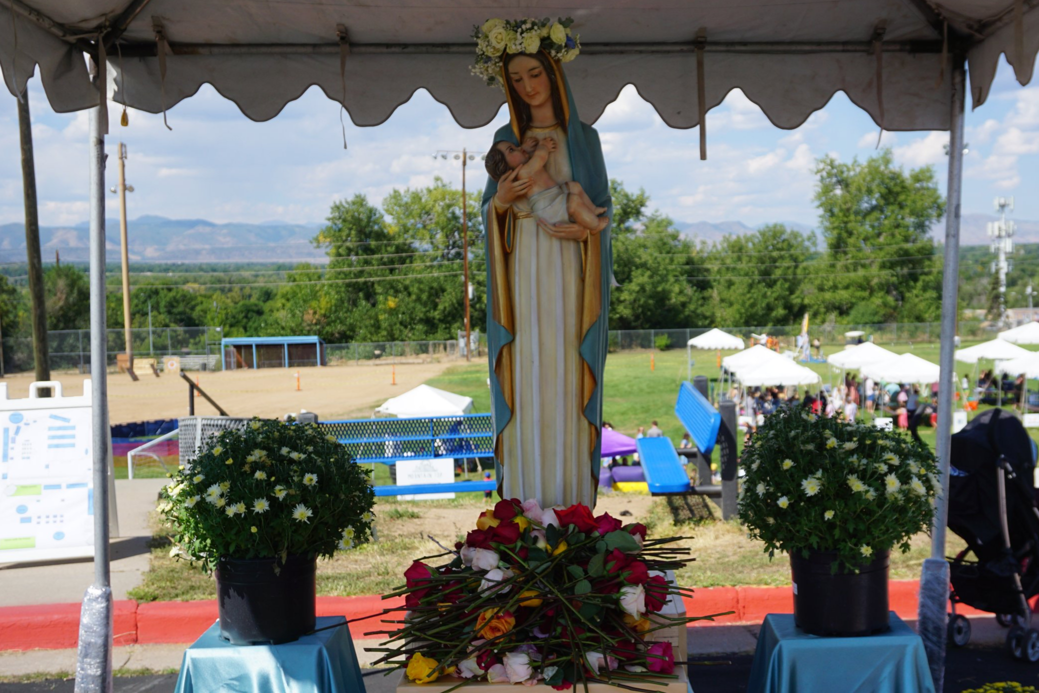 Outdoor religious shrine with a statue of the Virgin Mary holding baby Jesus, decorated with a flower crown, surrounded by flower arrangements, under a tent at a community event with trees, tents, and people in the background.
