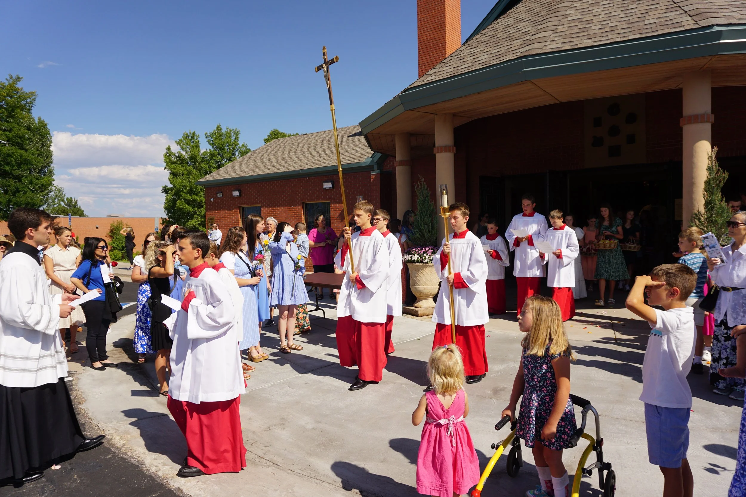 Group of children and teenagers gathered outside a church, with some dressed in choir robes. The scene appears to be part of a religious ceremony or event on a sunny day.